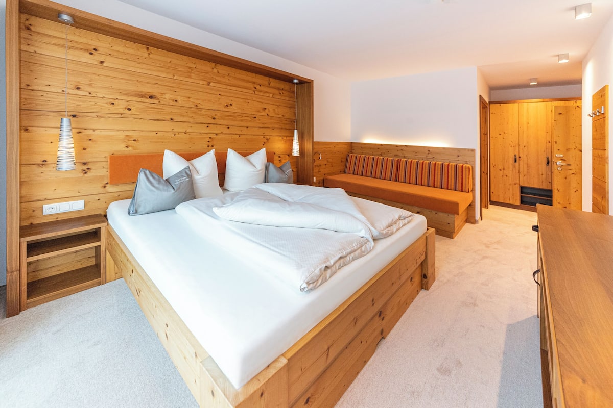 Uma cama de madeira com lençóis brancos, um banco com almofadas e um guarda-roupa de madeira.