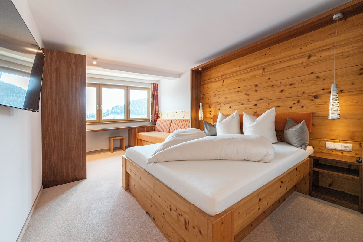 Quarto com cama de madeira, um banco, TV e janela com vista para as montanhas.