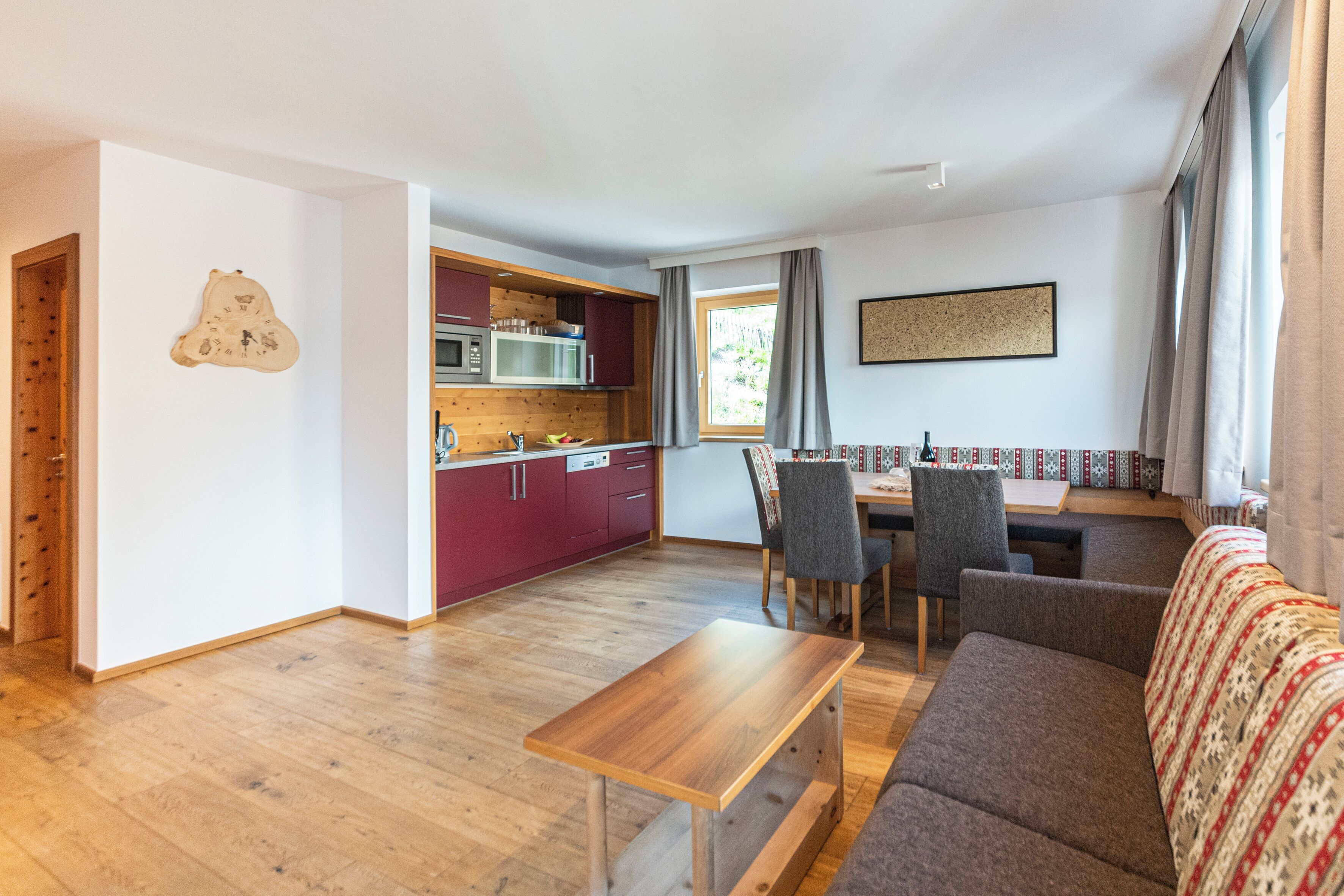 A compact living space with a wooden floor, a dining table with chairs, a sofa, a kitchen area with cabinets and appliances, and a window with curtains.