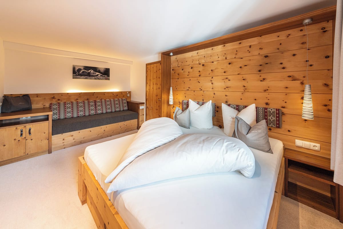 Quarto com cabeceira de madeira, cama com roupa de cama branca, um banquinho com almofadas estampadas e luminária fixada na parede.