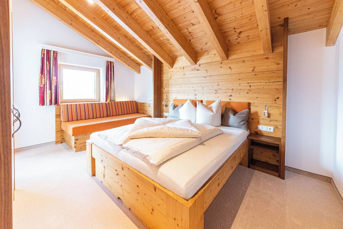 Quarto de madeira com cama, banco e janela.