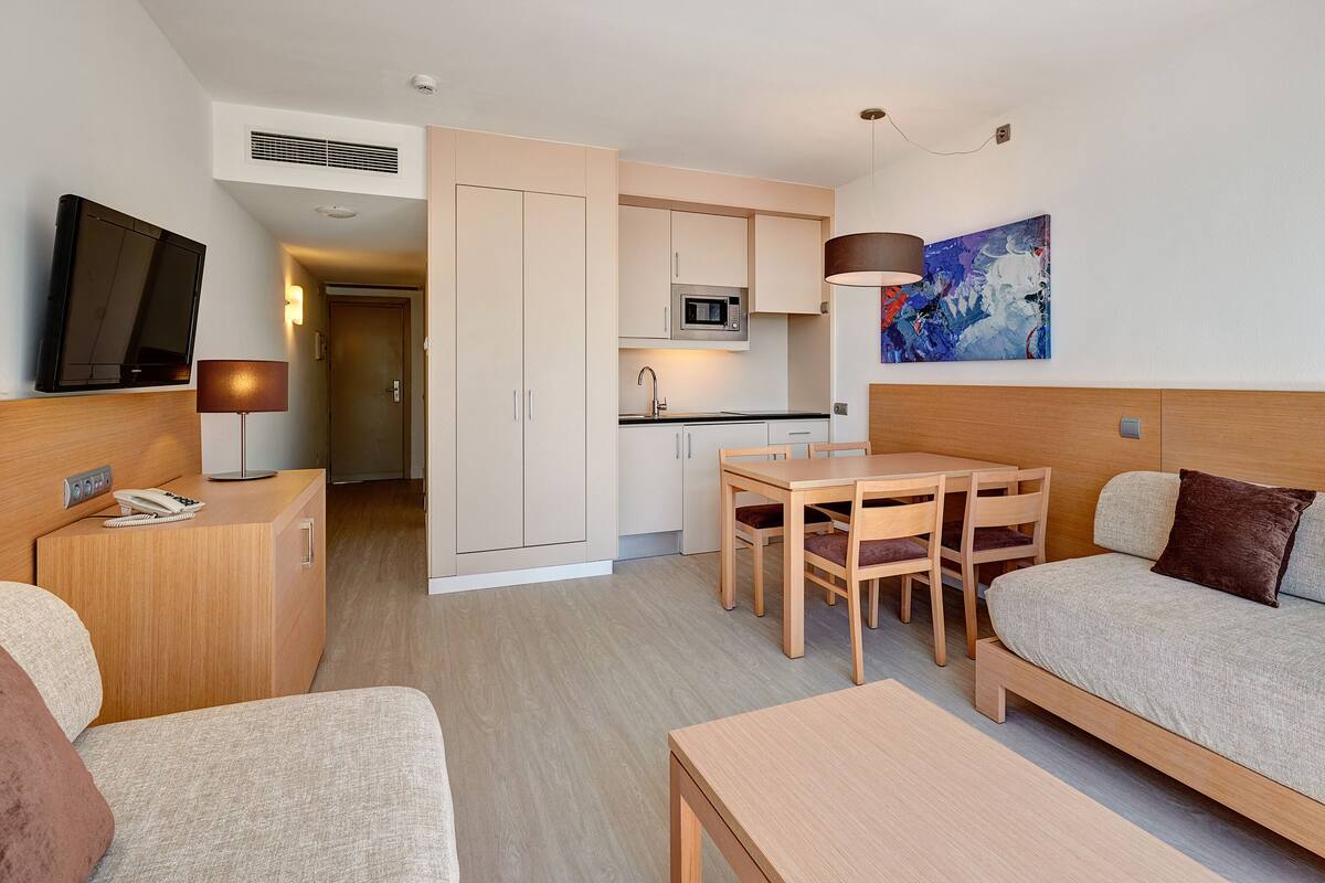 A compact hotel room with a kitchenette, a dining area, a sofa, and a TV.