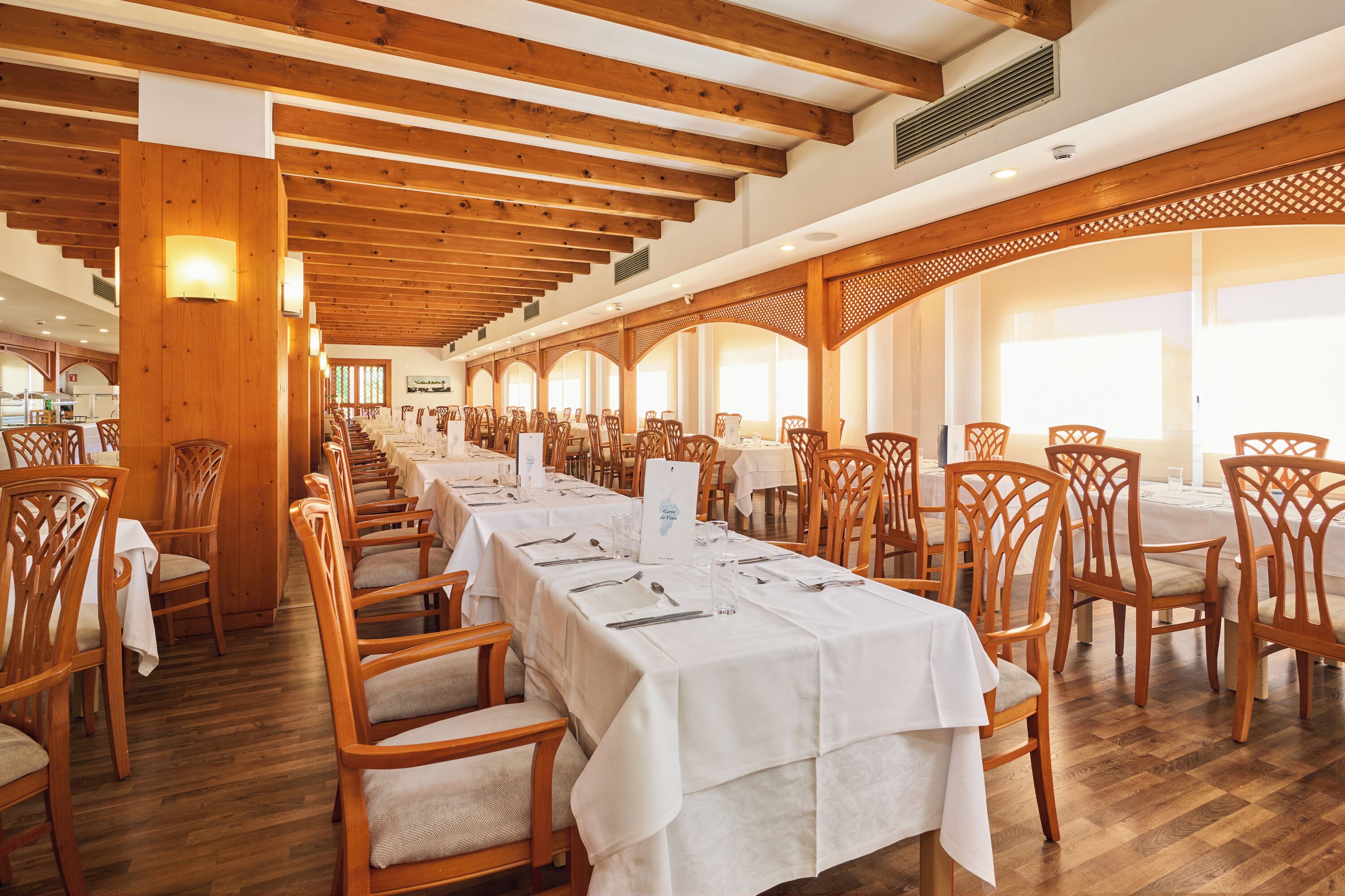 A long dining table with white tablecloths and wooden chairs in a spacious room with wooden beams on the ceiling.