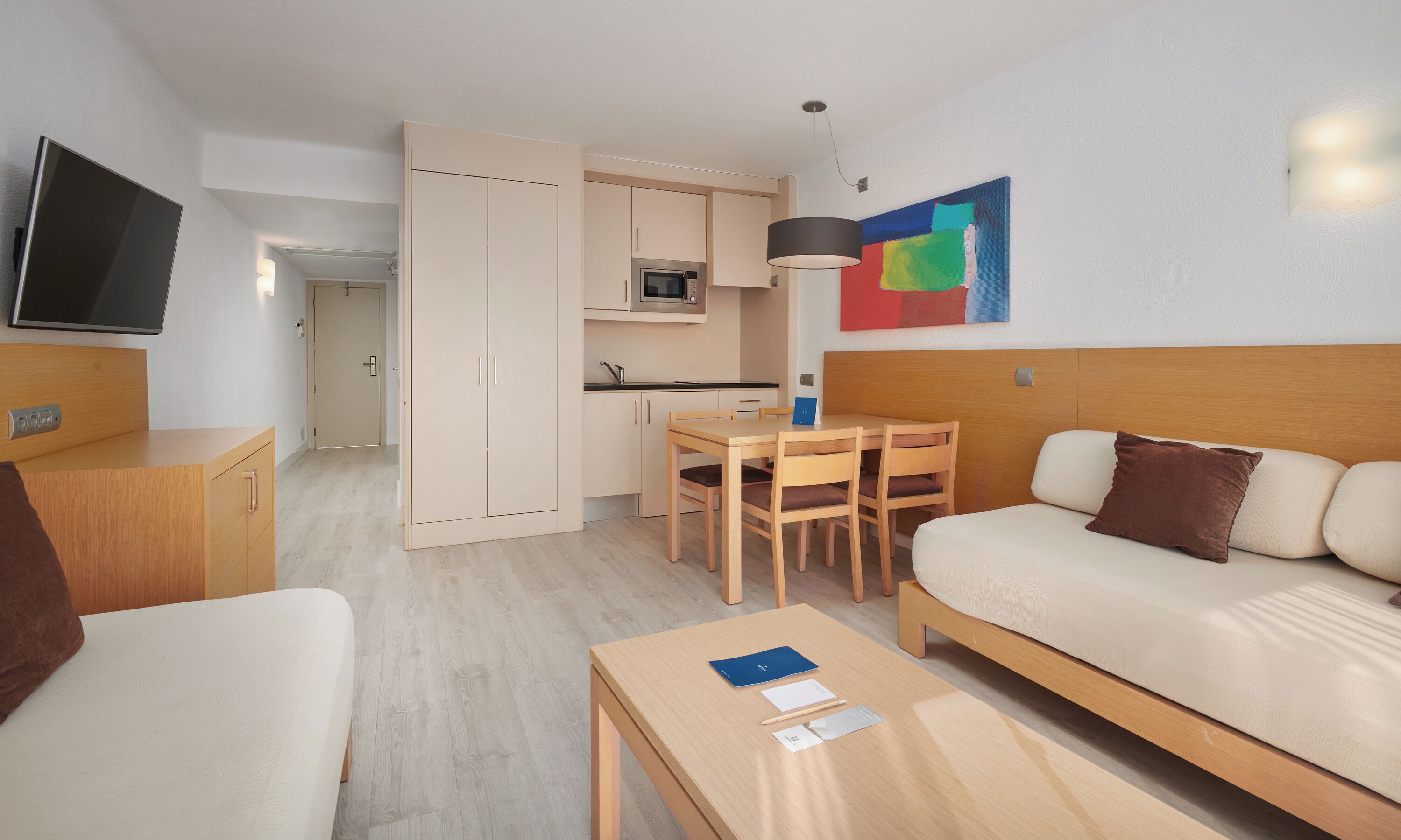 A modern hotel room with a sofa, a small table, a TV, a kitchenette, and a painting on the wall.