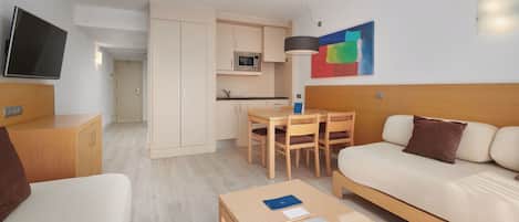 A modern hotel room with a sofa, a small table, a TV, a kitchenette, and a painting on the wall.