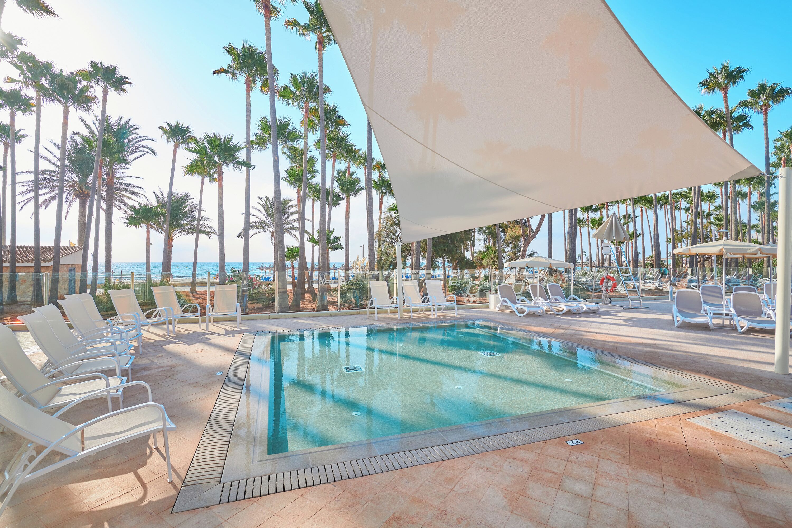 A pool area with lounge chairs and a shaded canopy, surrounded by palm trees and a view of the ocean.