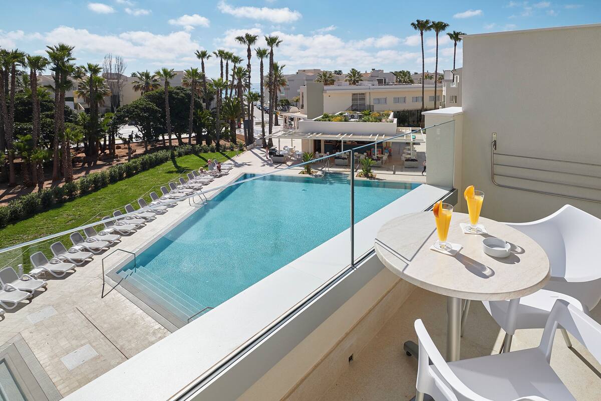 A rooftop pool with lounge chairs and a table with drinks, surrounded by palm trees and buildings.