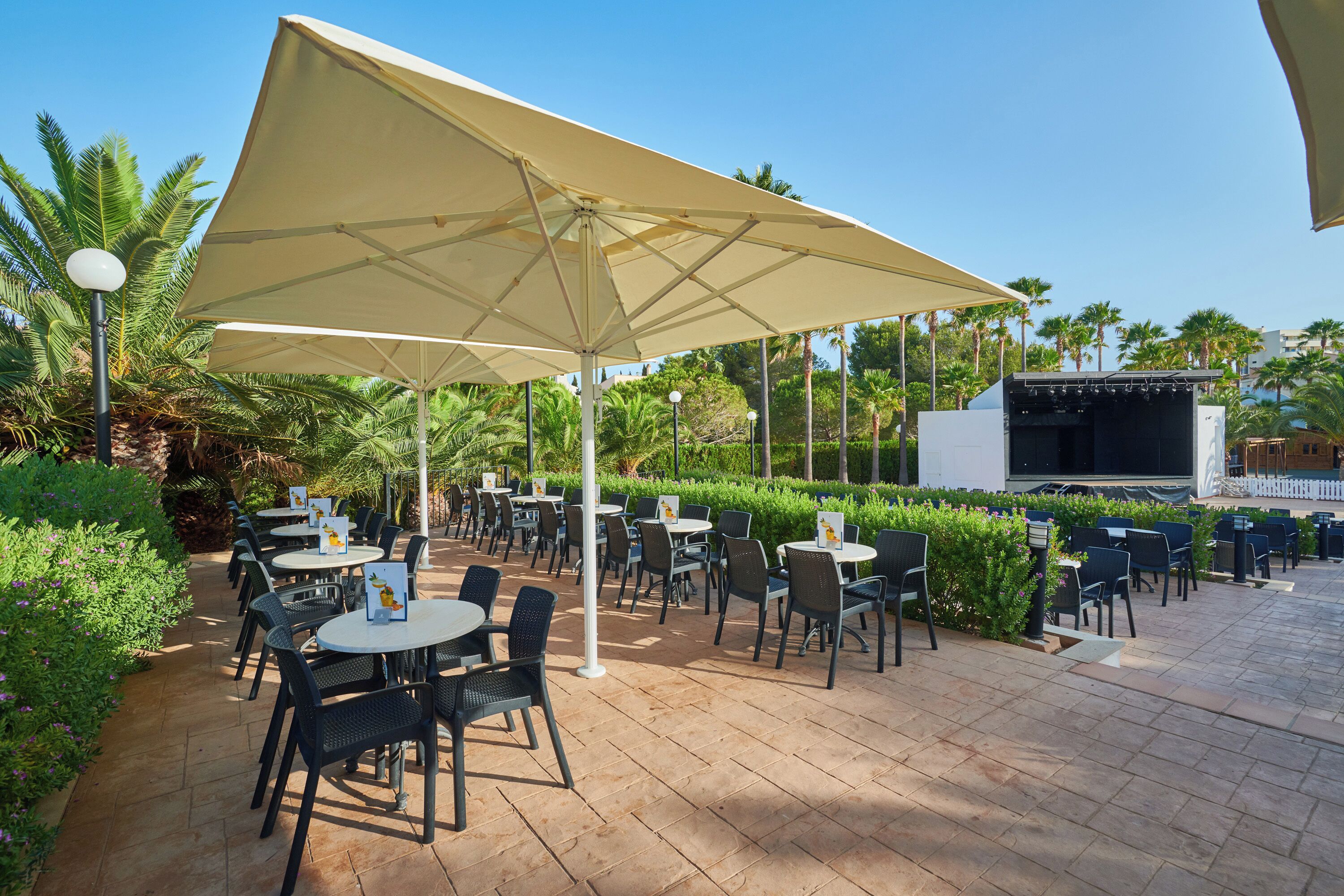 An outdoor patio area with round tables and chairs, covered by large umbrellas, surrounded by greenery and palm trees.