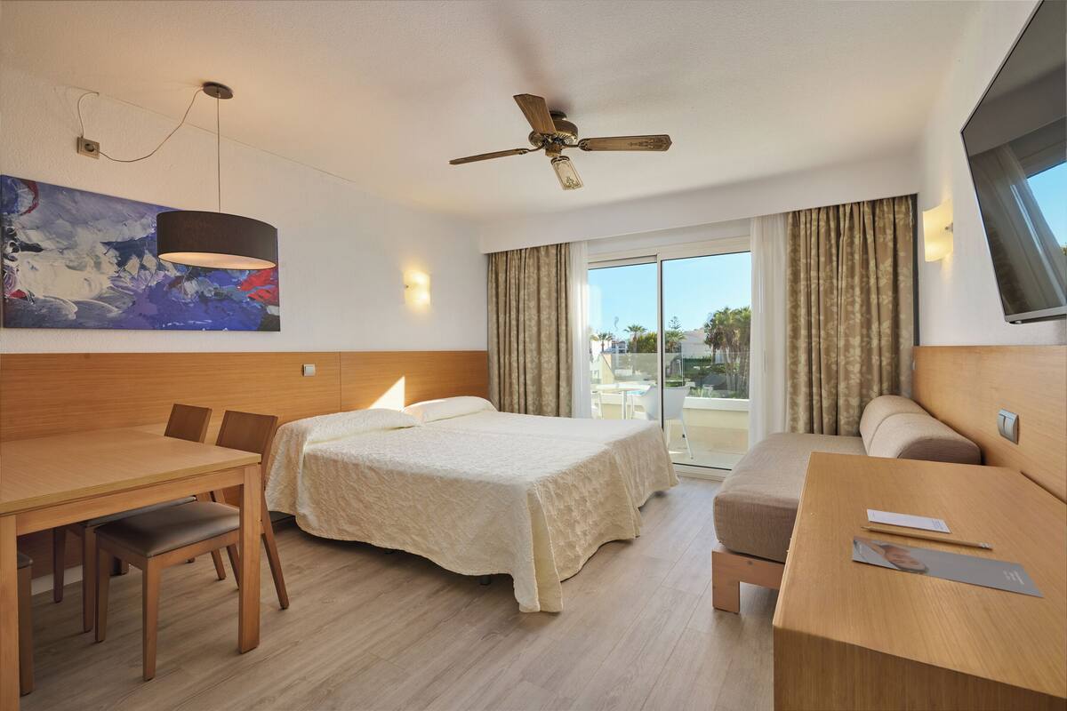 A hotel room with a bed, a desk, a chair, a table, a ceiling fan, a painting, and a window with curtains.