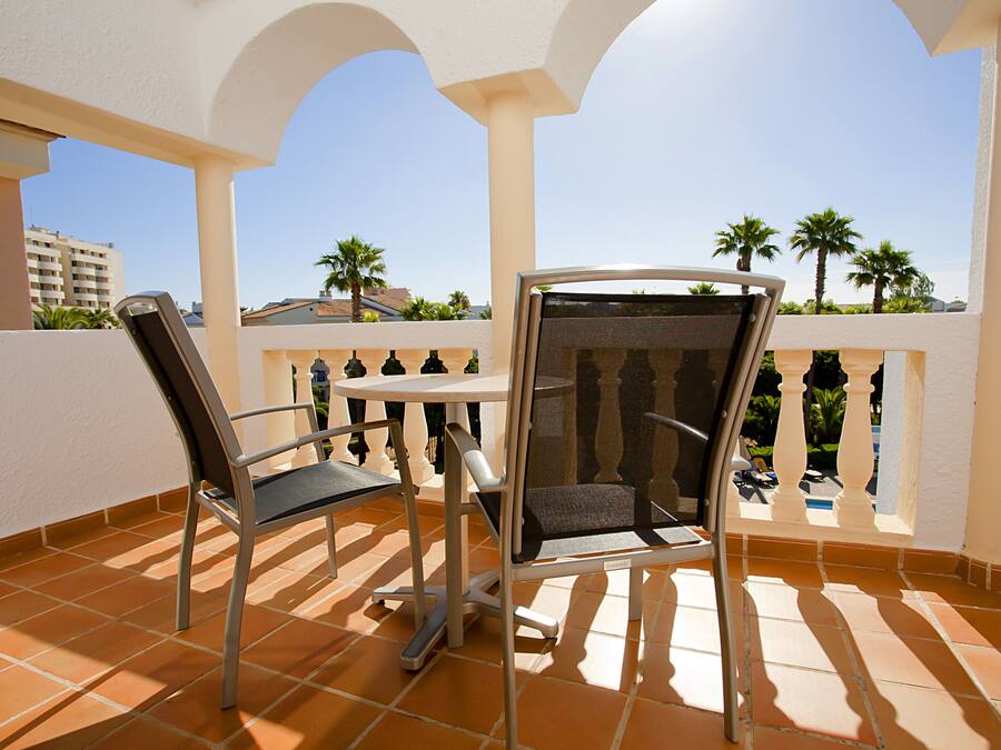 A terrace with two chairs and a table, overlooking a pool and palm trees.