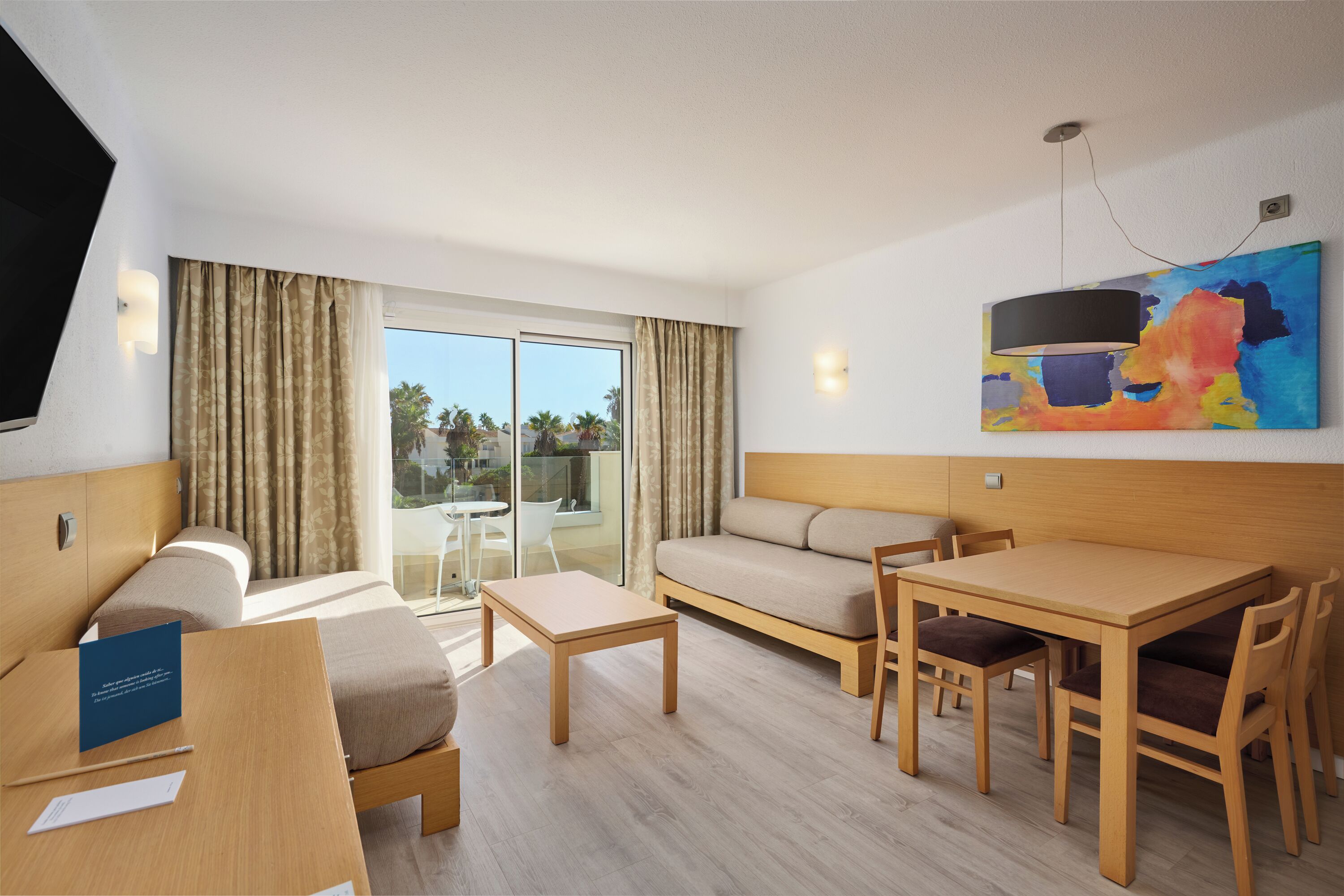 A modern hotel room with a bed, a sofa, a dining table with chairs, a TV, and a large window with curtains.
