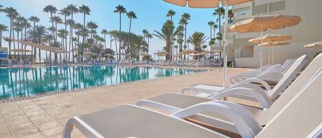 A poolside area with lounge chairs and umbrellas, palm trees in the background, and a clear sky.