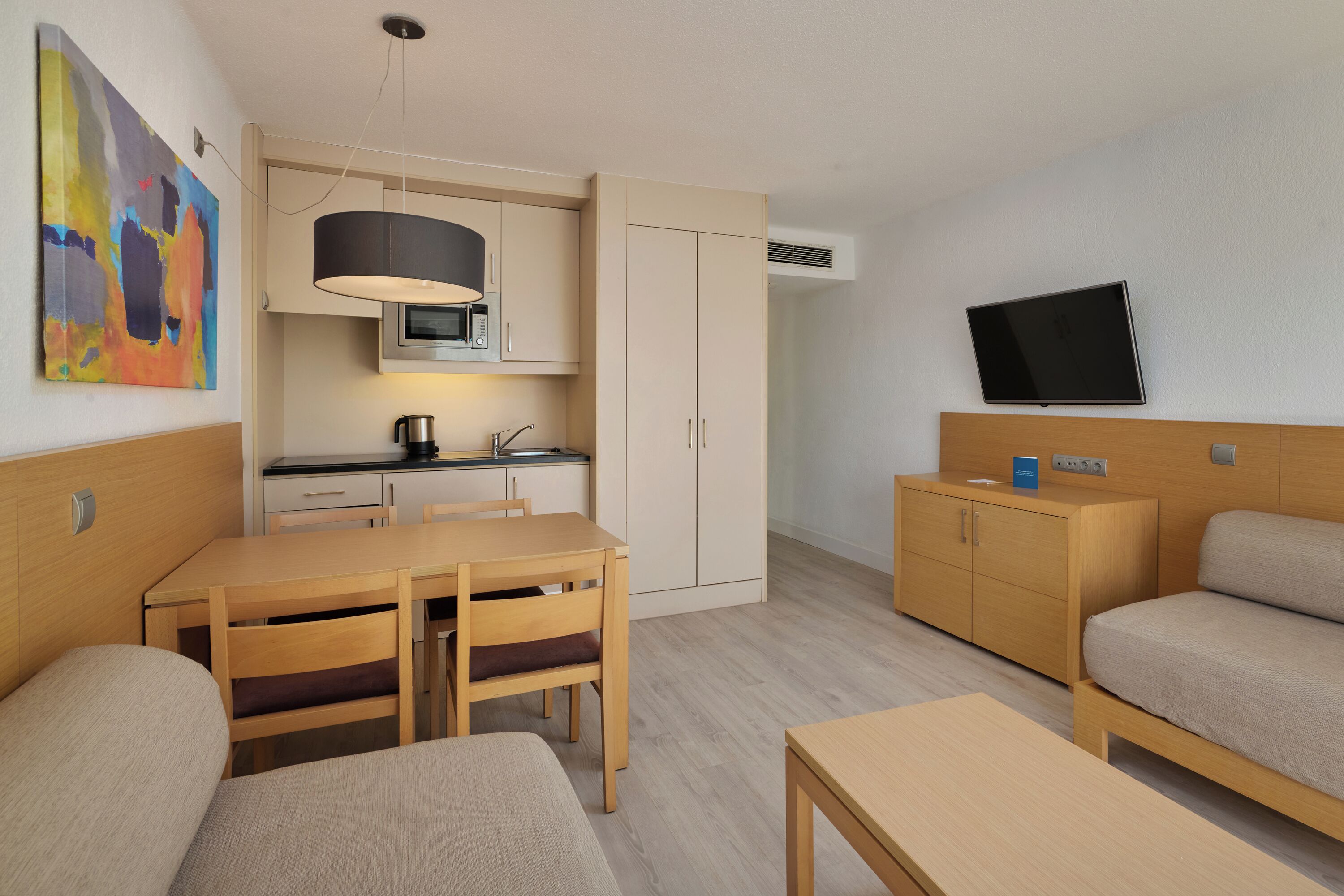 Una habitación de hotel moderna con kitchenette, comedor, sofá y televisor montado en la pared.