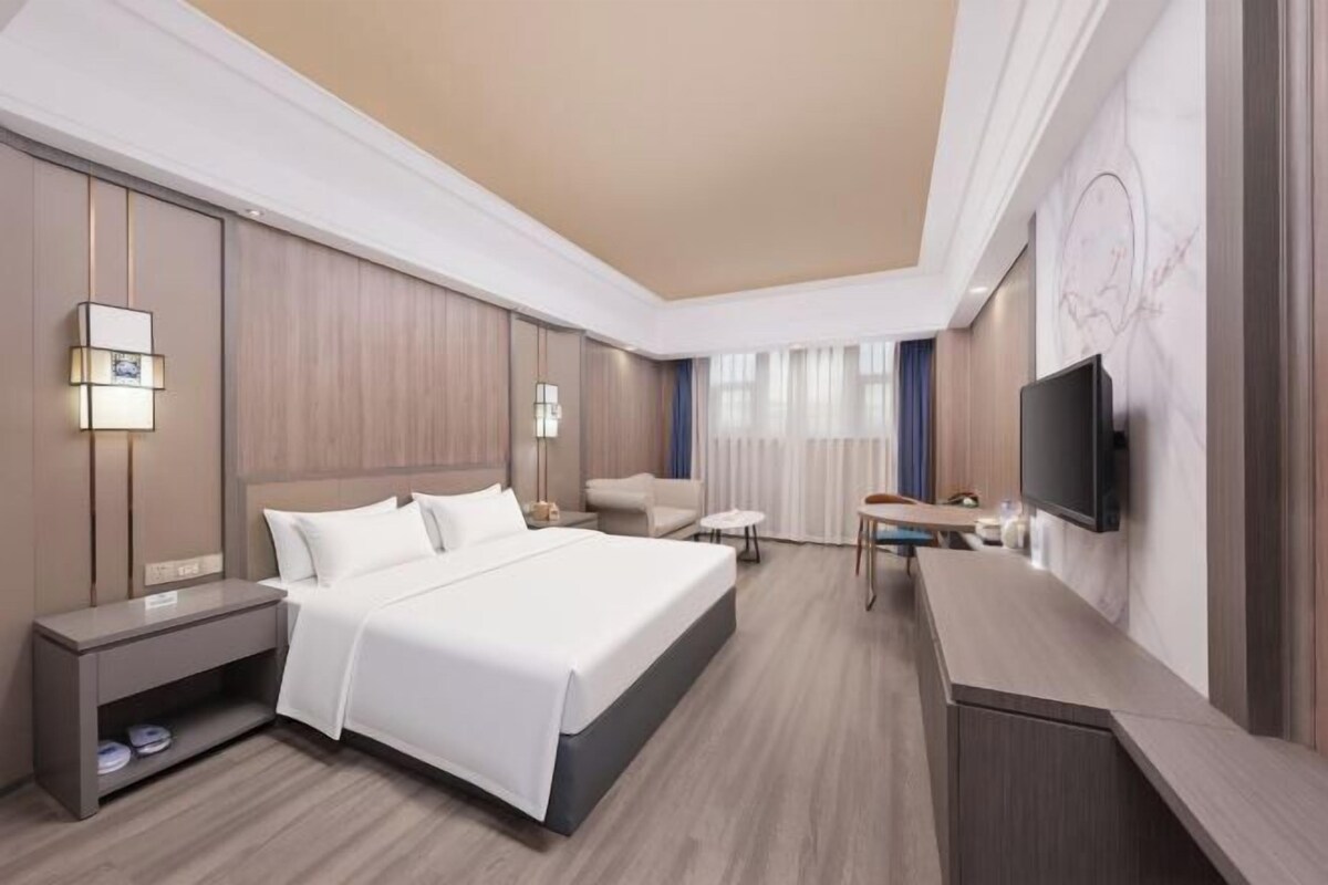 A modern hotel room with a large bed, a desk, a chair, a TV, and a nightstand.