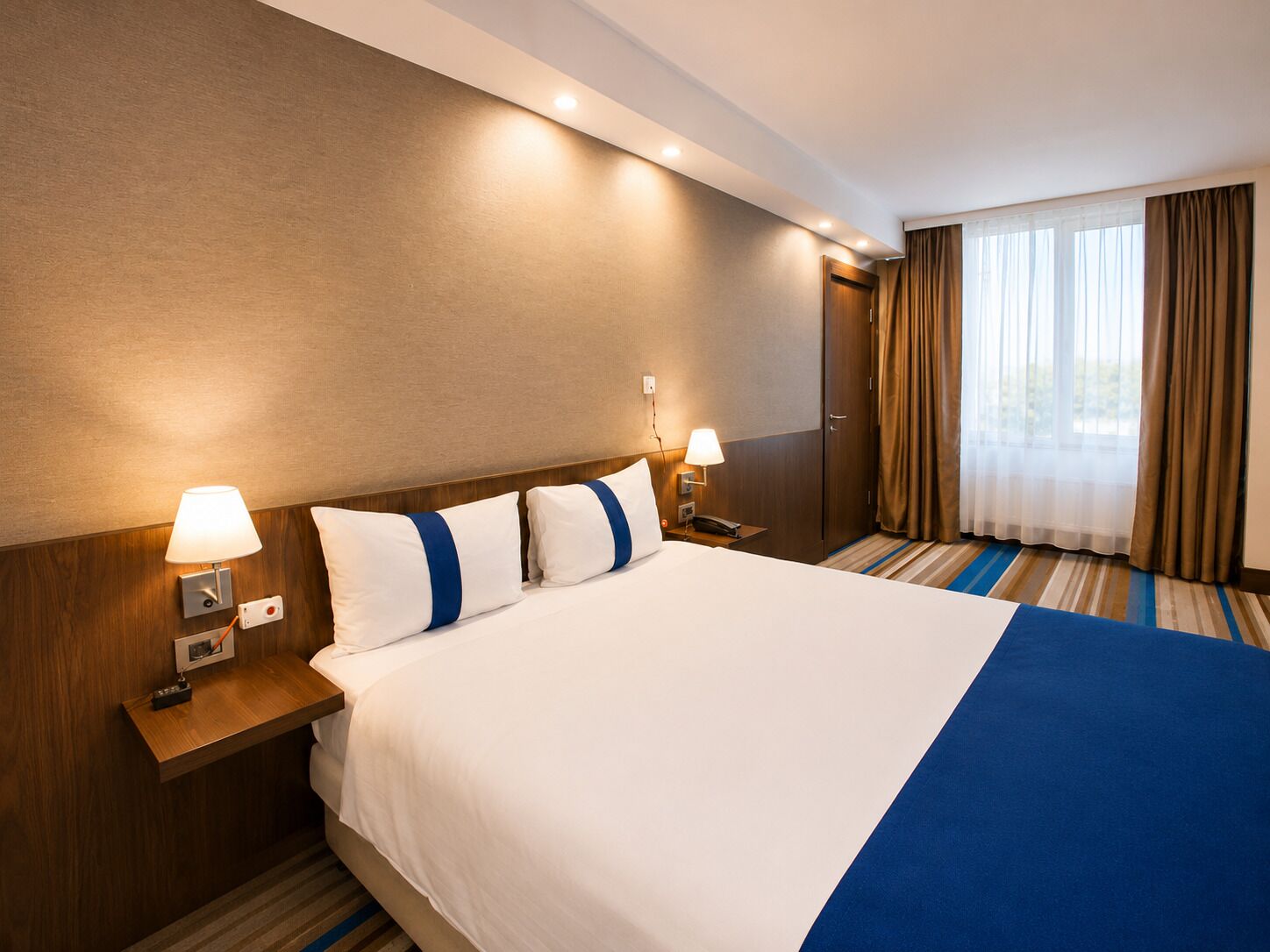 A hotel room with a large bed, two bedside lamps, a wooden headboard, a window with curtains, and a striped carpet.