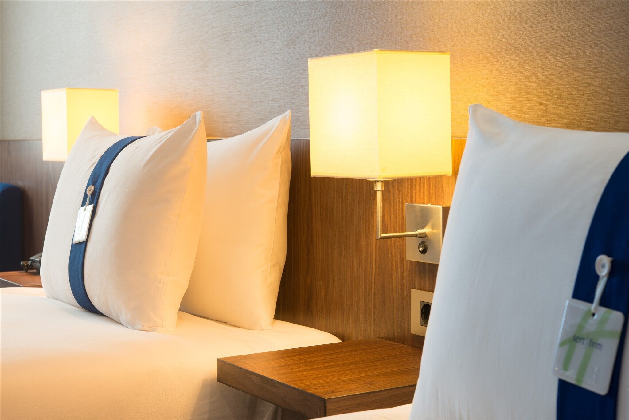 A hotel room with a bed, pillows, a nightstand, and a lamp.