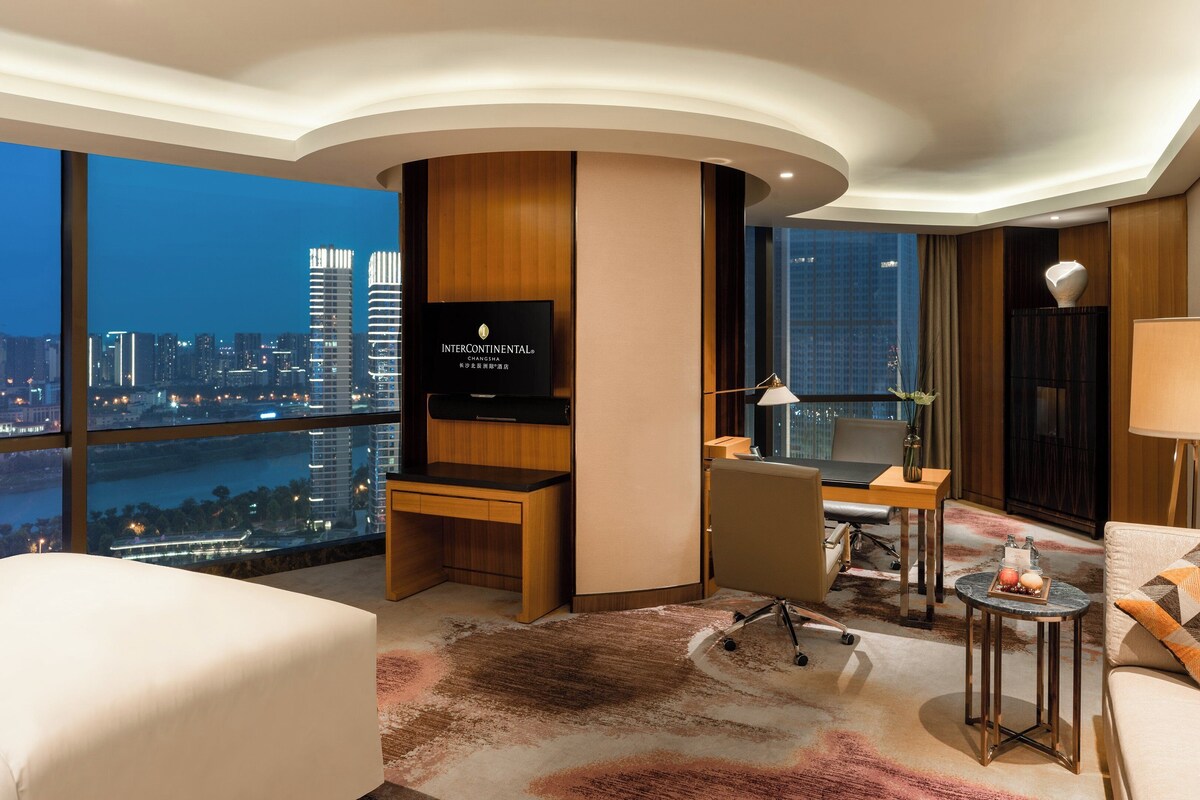 A modern hotel room with a city view, a desk, a sofa, and a TV.