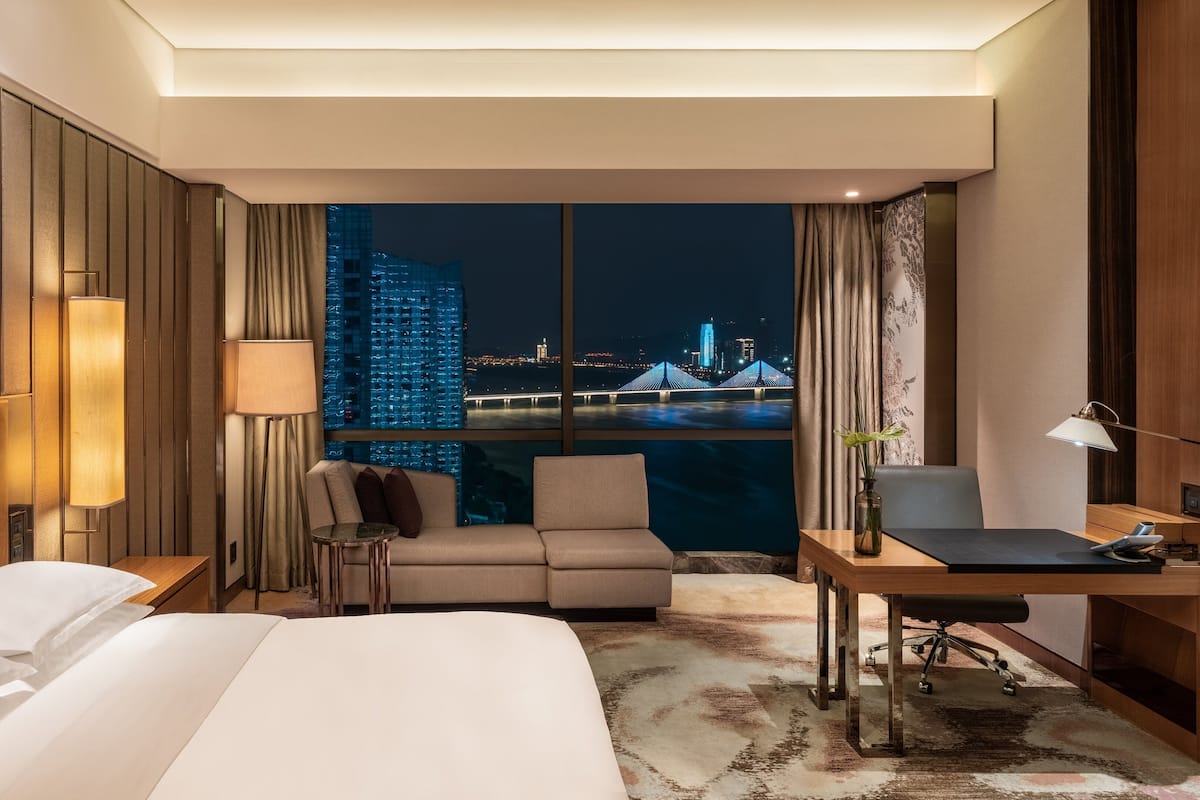 A hotel room with a bed, a desk, a chair, a sofa, and a view of a cityscape at night.