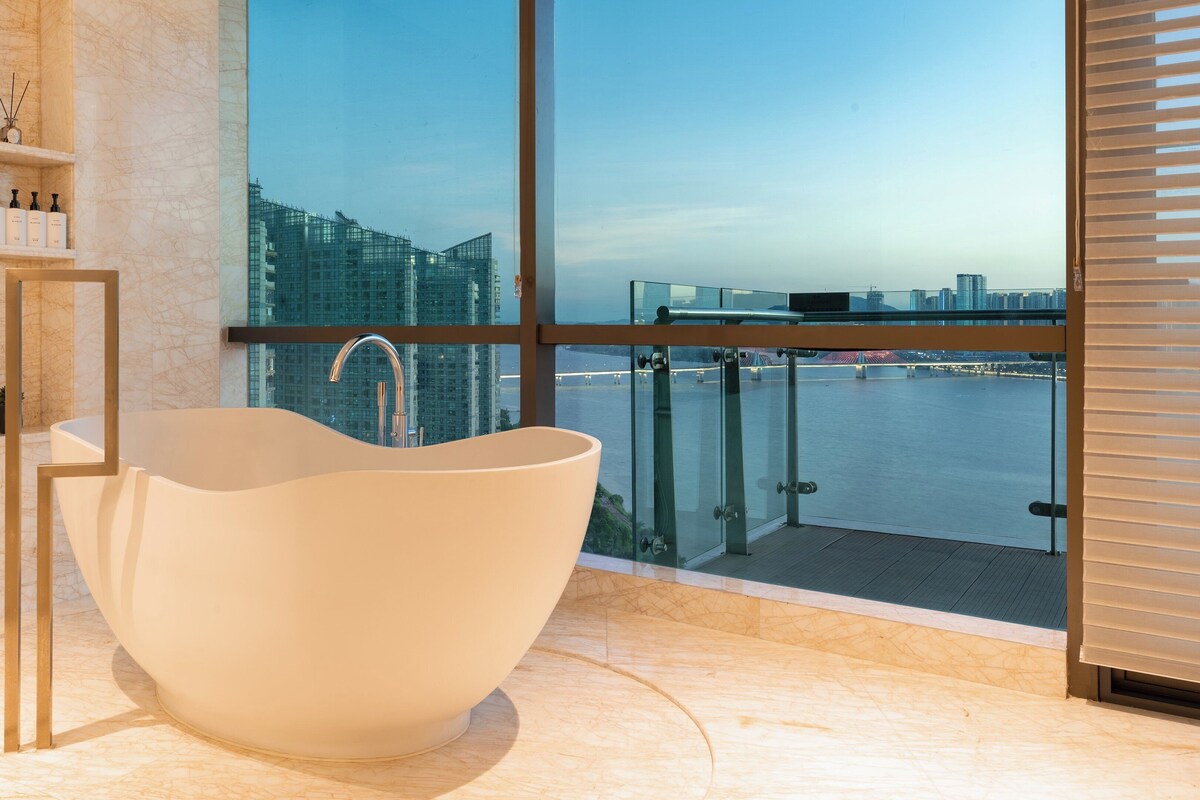 A modern bathroom with a freestanding bathtub, marble walls, and a city view through large windows.
