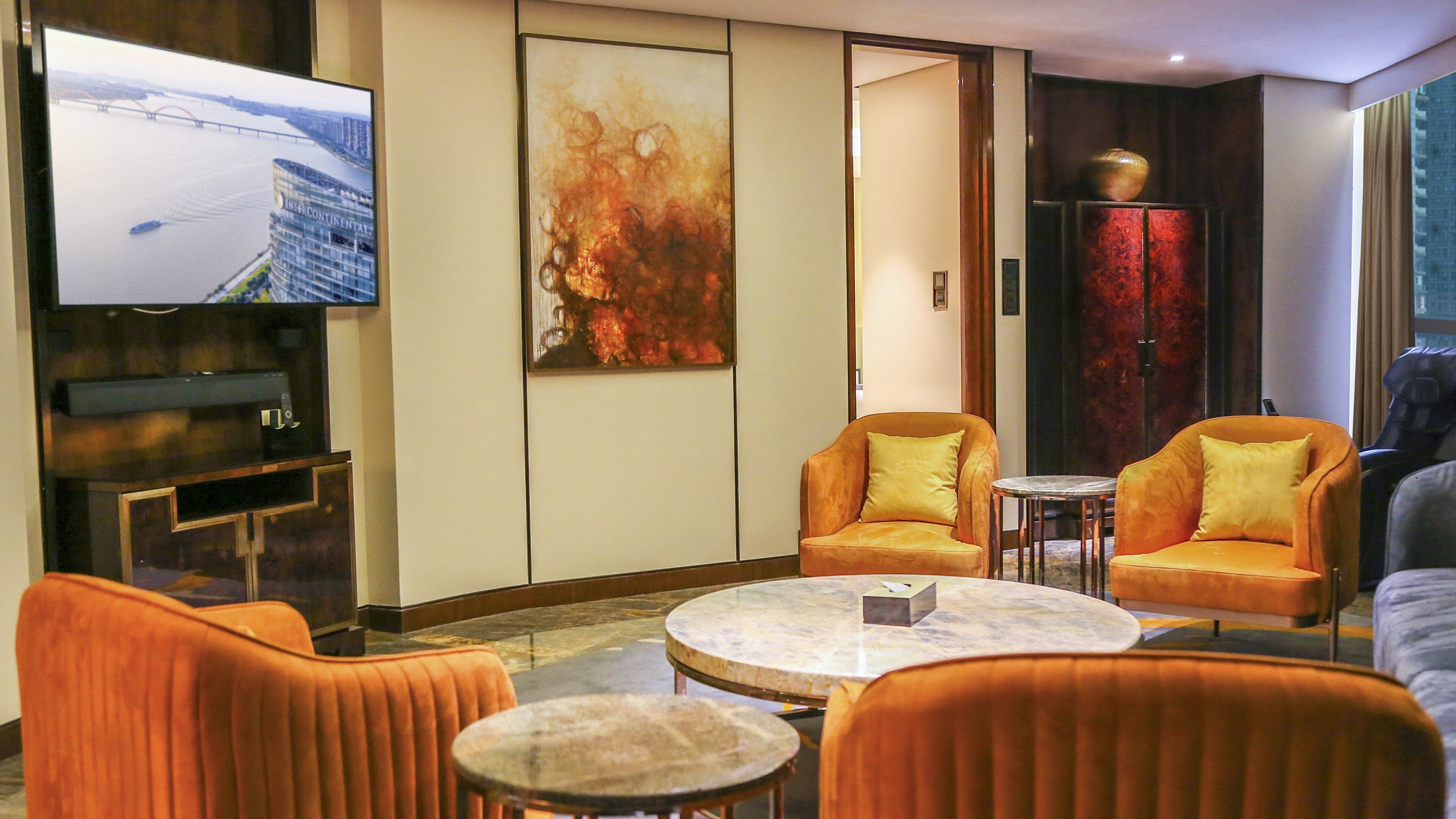 Een hotelkamer met een grote flatscreen televisie, oranje fauteuils, een marmeren salontafel en een groot abstract schilderij.