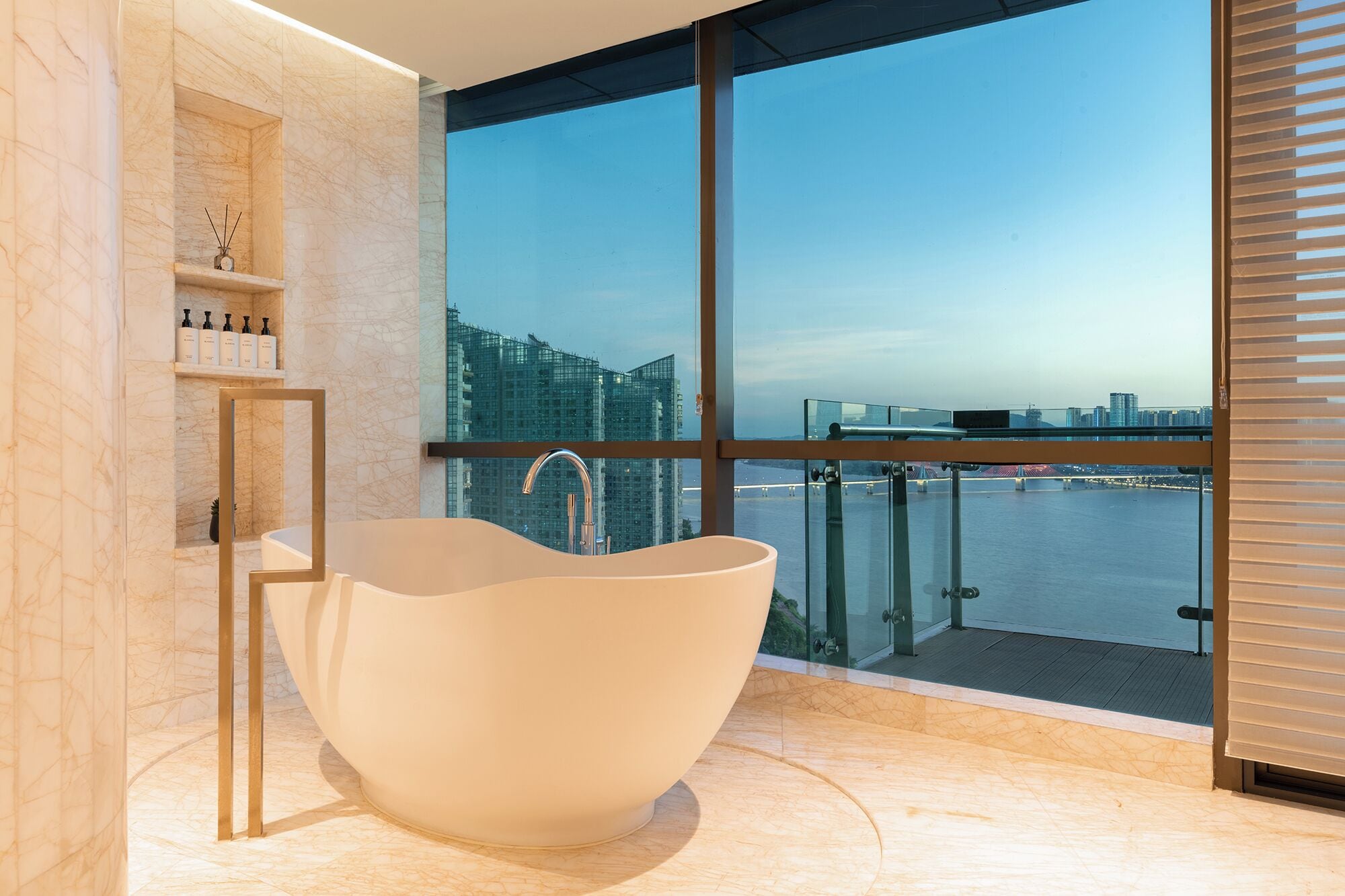A modern bathroom with a freestanding bathtub, marble walls, and a large window offering a city view.