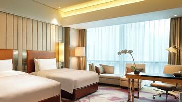 A hotel room with two beds, a desk, a chair, a sofa, and a large window with curtains.