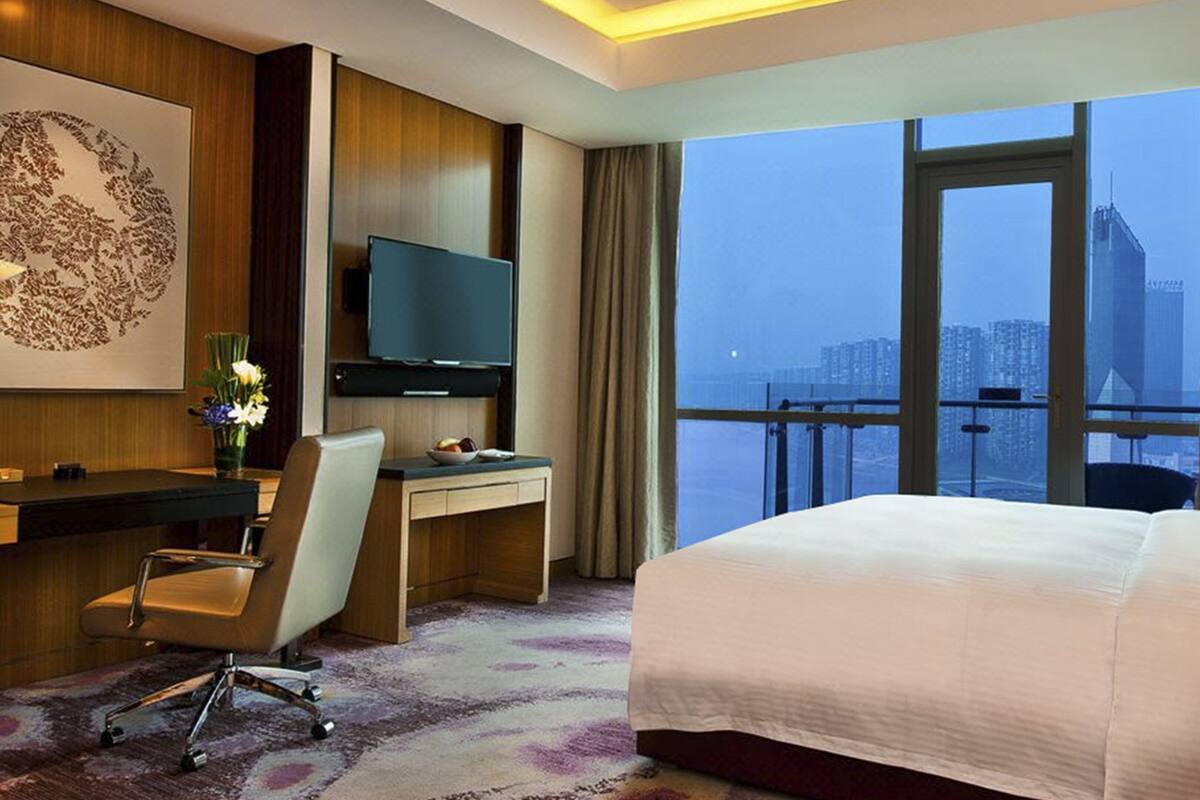 A hotel room with a large bed, a desk with a chair, a television, and a view of a cityscape through large windows.
