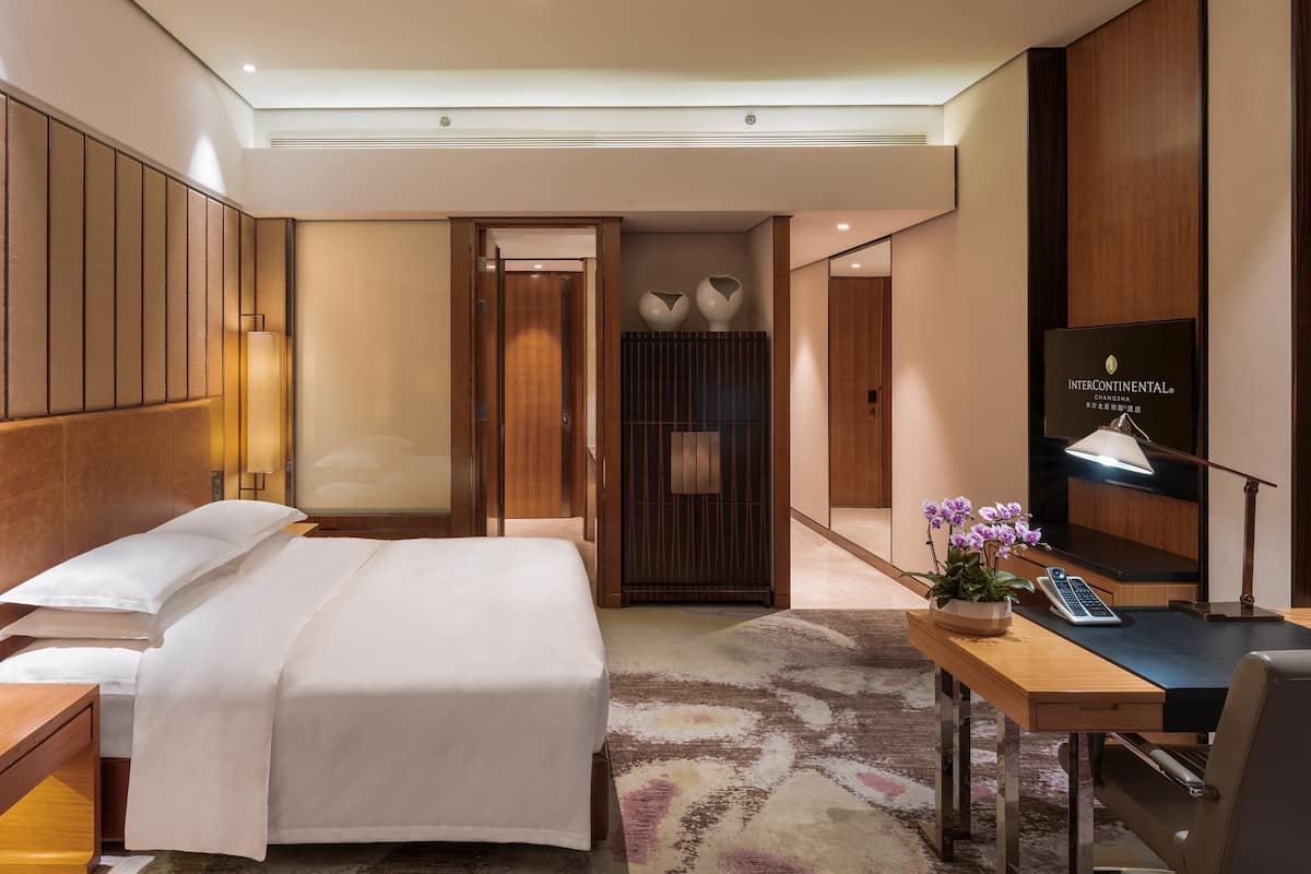 A hotel room with a bed, bedside table, lamp, desk with a phone, and a TV displaying 'INTERCONTINENTAL CHANGSHA'.