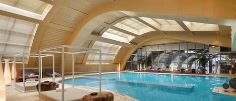 Una piscina interior con vistas despejadas al cielo a través de amplios ventanales, un área de descanso con asientos y un elemento decorativo de madera.