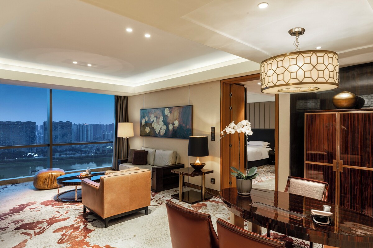 A modern hotel room with a large window offering a city view, a comfortable seating area, a dining table, and a bed with a wooden wardrobe.