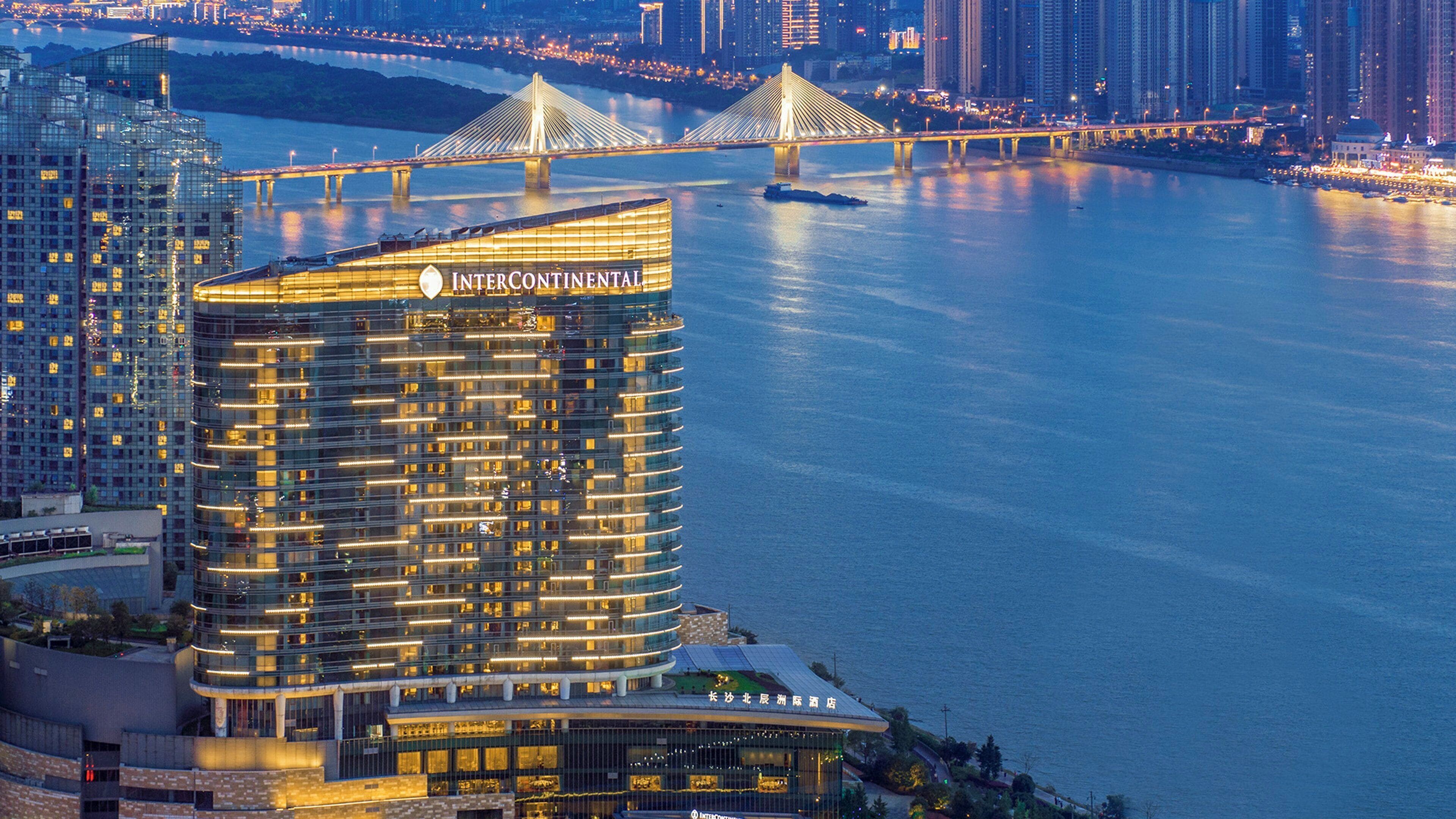 A high-rise hotel with a modern design, located near a river with a bridge in the background.