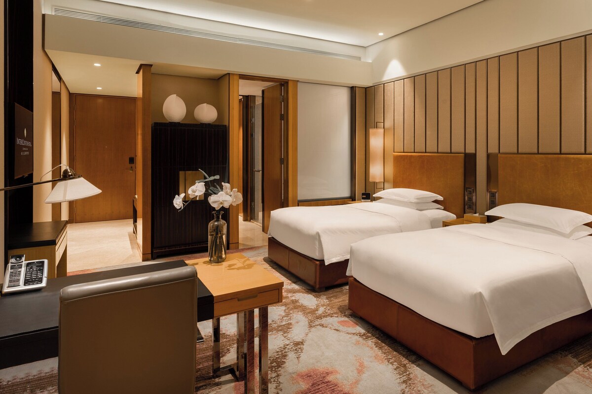 A hotel room with two beds, a desk, a chair, a lamp, and a vase with flowers.