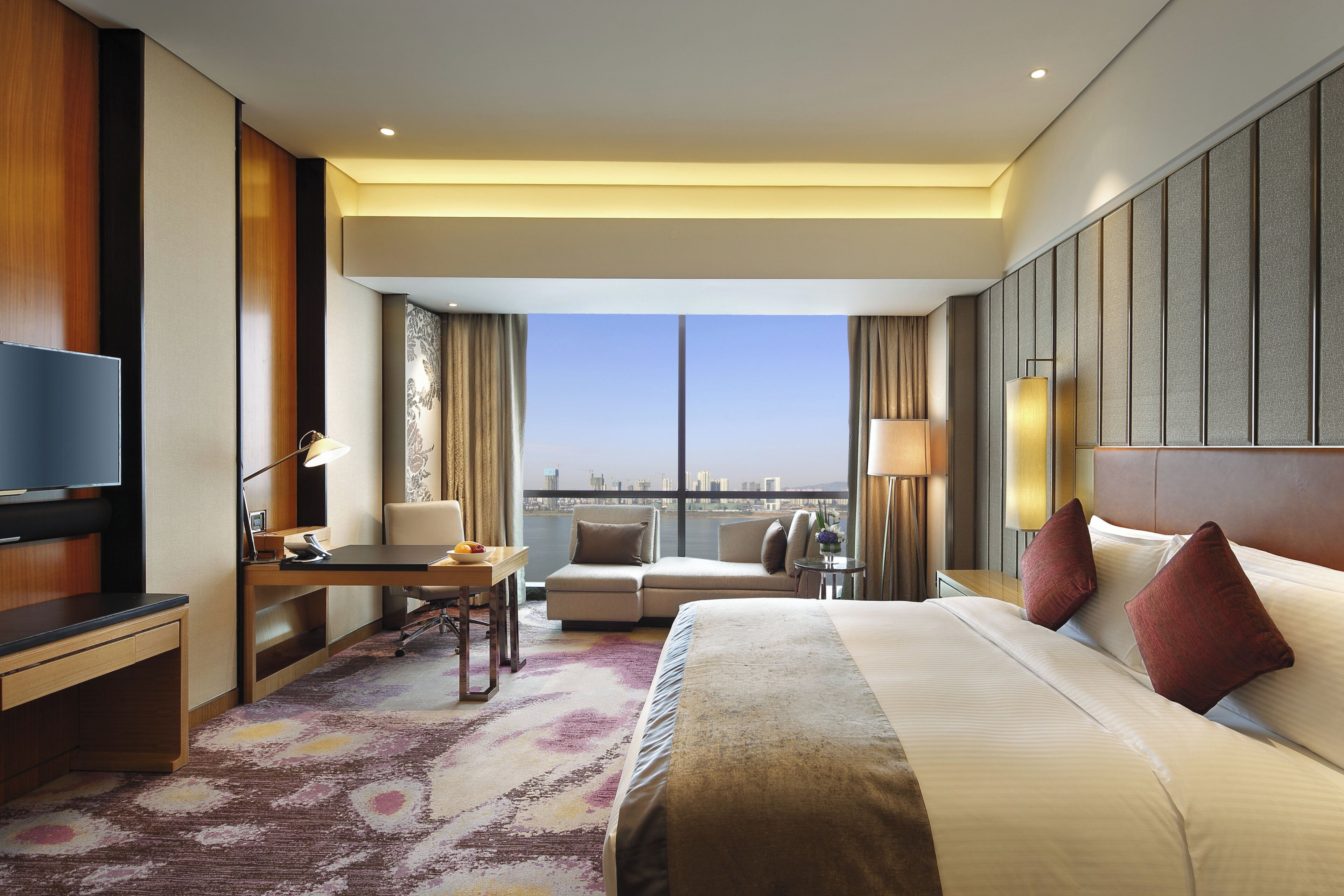 Ein Hotelzimmer mit einem Bett, einem Schreibtisch, einem Sessel sowie einem Fernseher und Blick auf die Stadt.