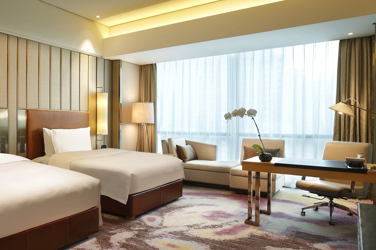 A hotel room with two beds, a desk, a chair, a lamp, and a view of the city through large windows.