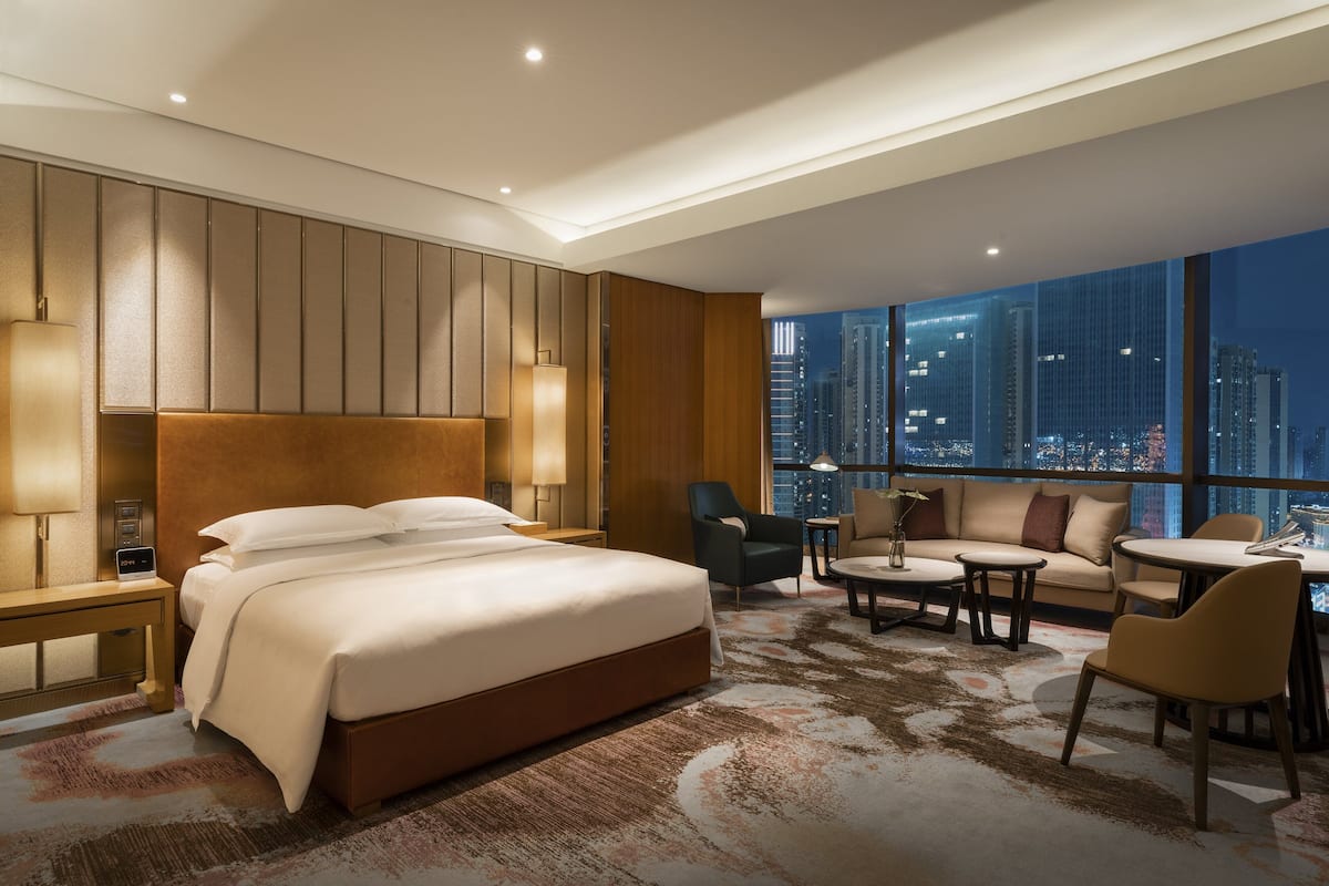 A modern hotel room with a large bed, a sofa, a dining table, and a city view.