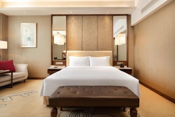 A hotel room with a large bed, a bedside table, a nightstand, a red pillow, a white wall, and a framed picture.