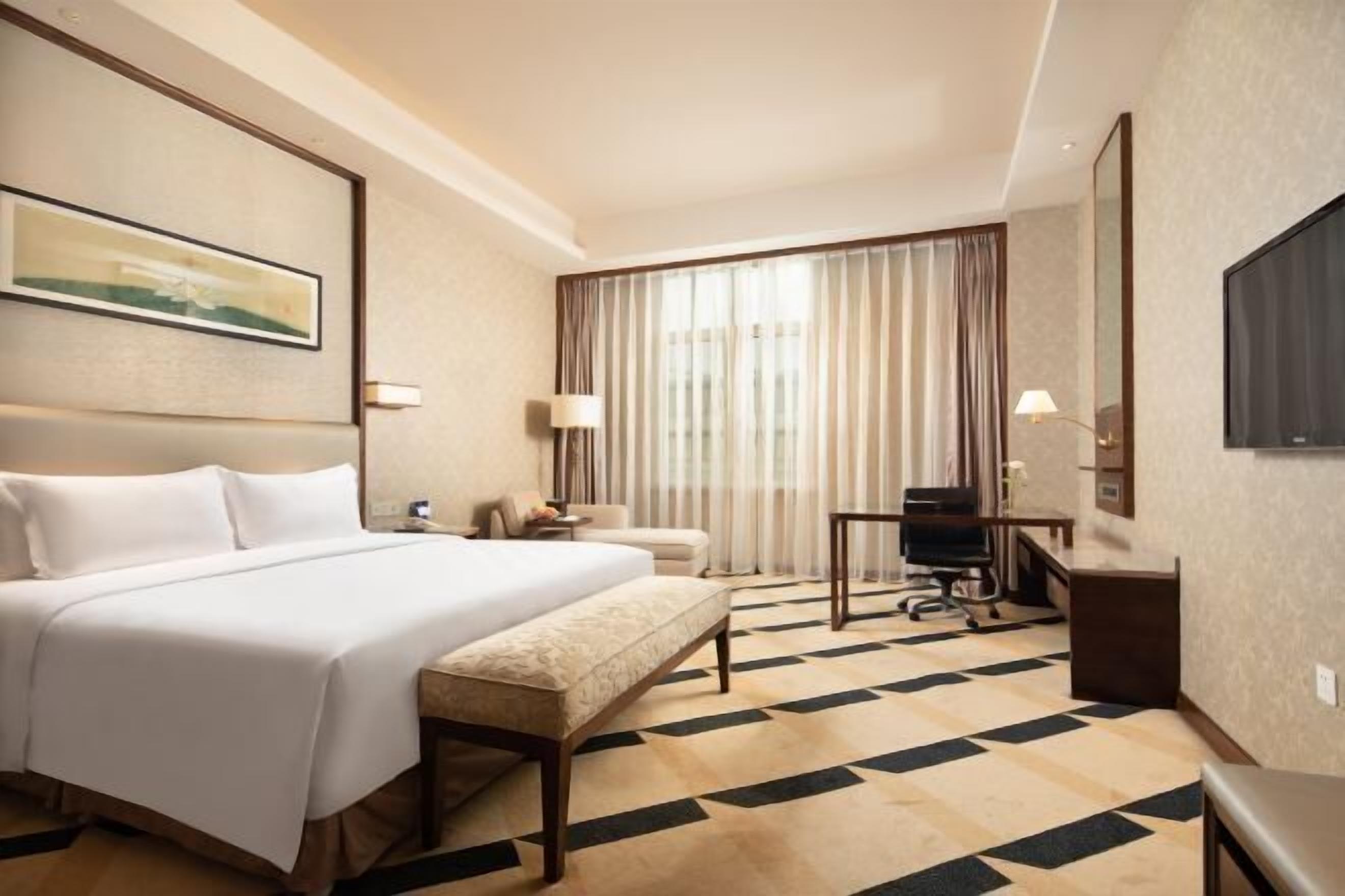 A hotel room with a large bed, a desk, a chair, a bench, a TV, and a window with curtains.