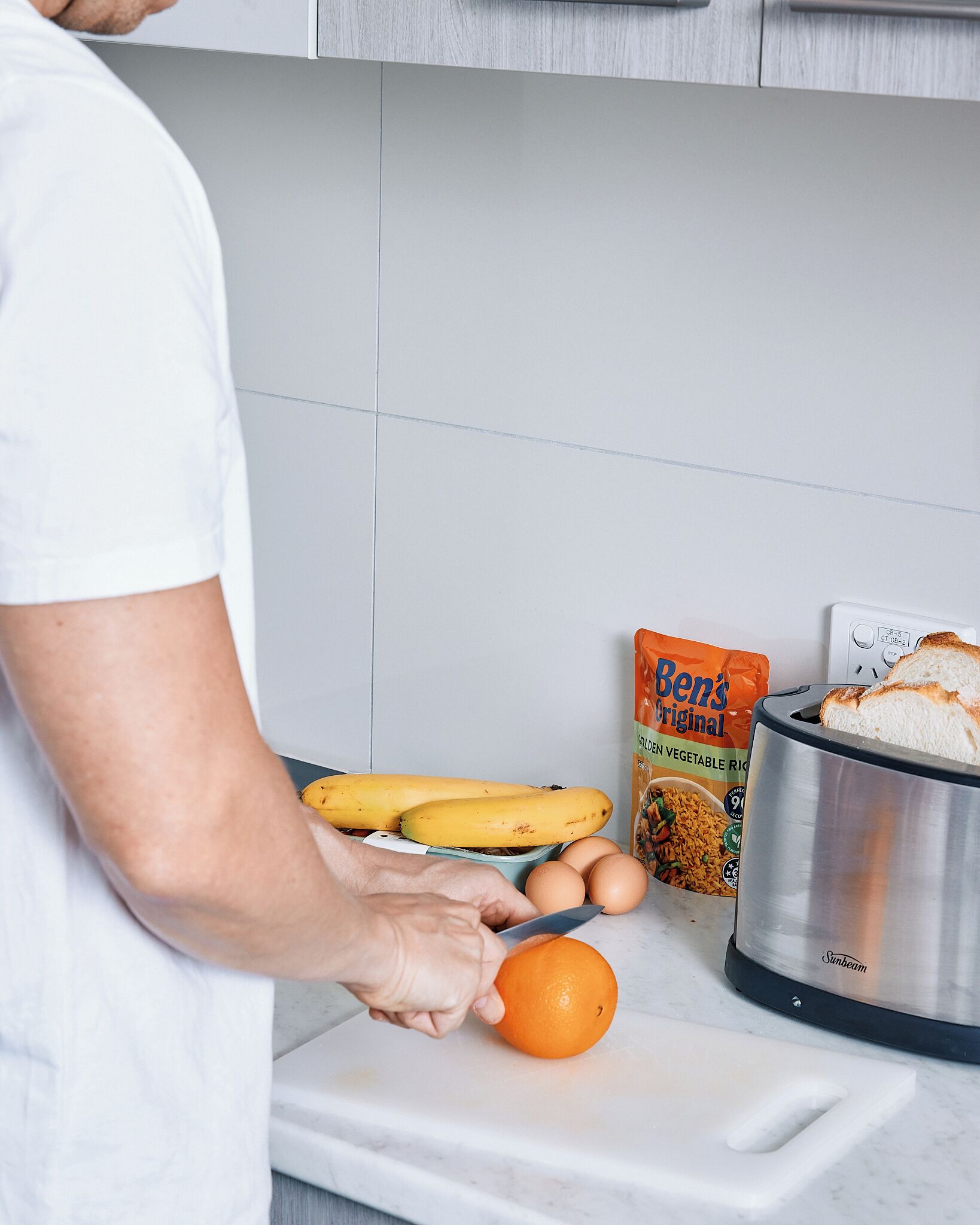 Iemand bereidt eten met bananen, eieren en een sinaasappel op een aanrecht.