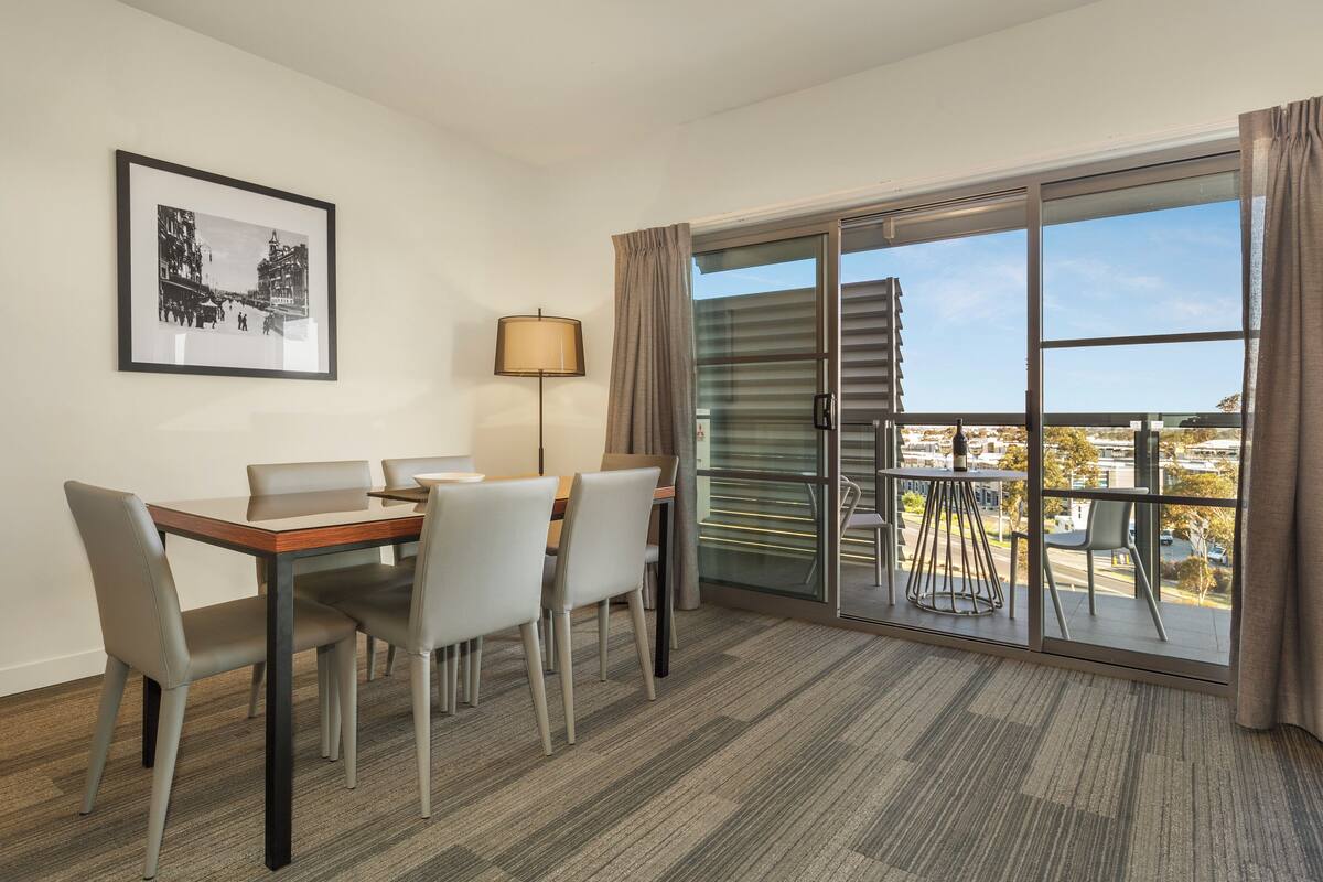 Ein Hotelzimmer mit Esstisch, Stühlen, einer Lampe, einem gerahmten Bild an der Wand und einem Balkon mit Aussicht.