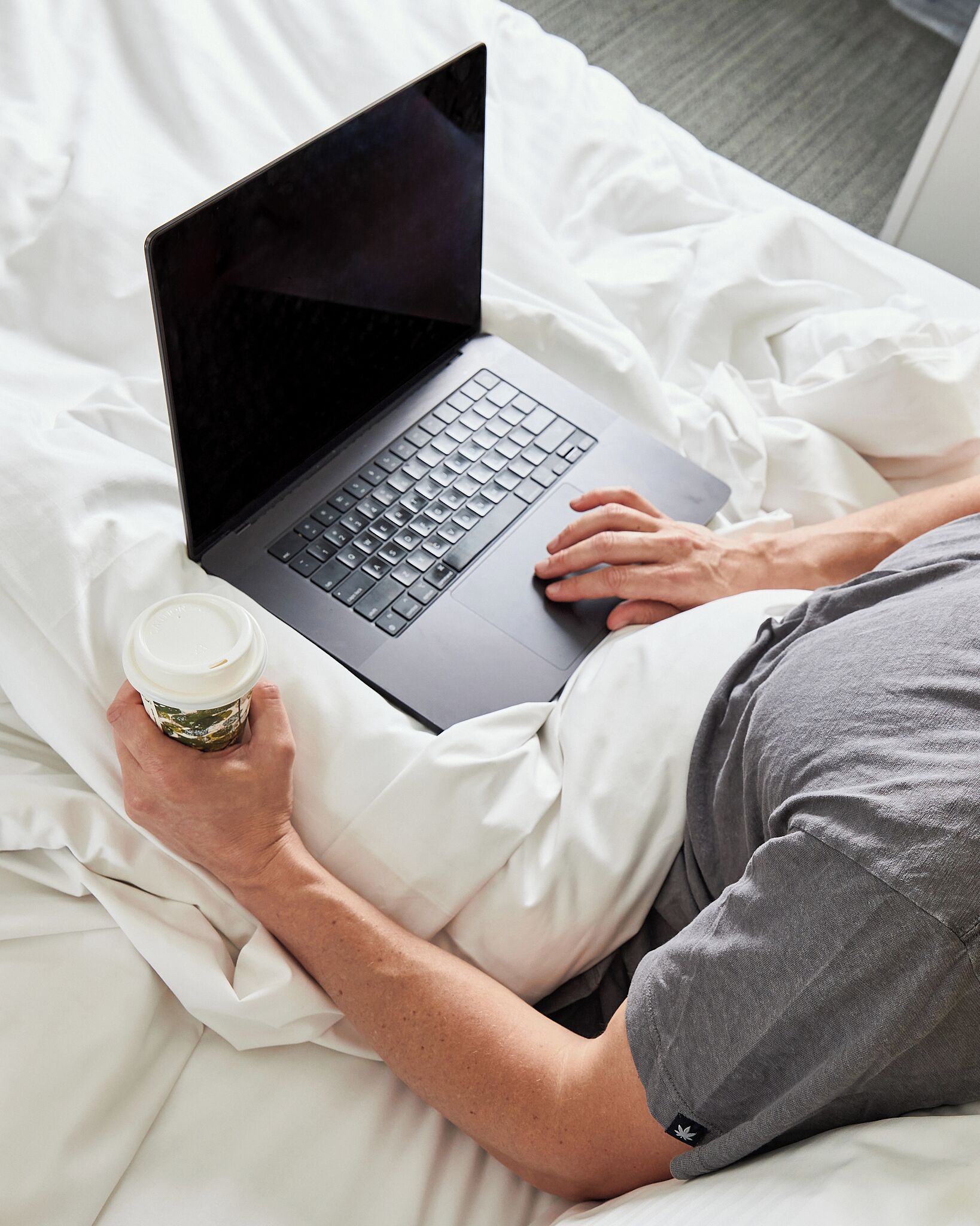 Una persona acostada en una cama con un ordenador portátil y una taza de café.