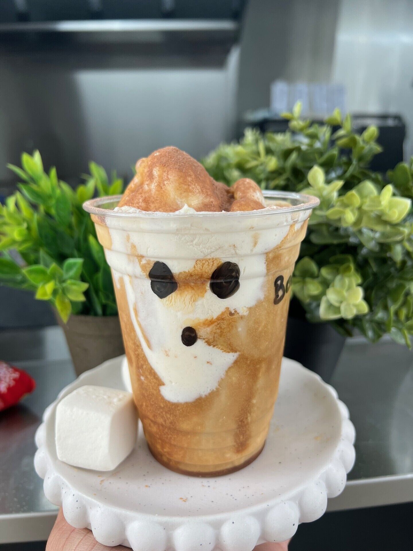 A cup of bubble tea with a decorative face on the side, placed on a white coaster with a small plant in the background.