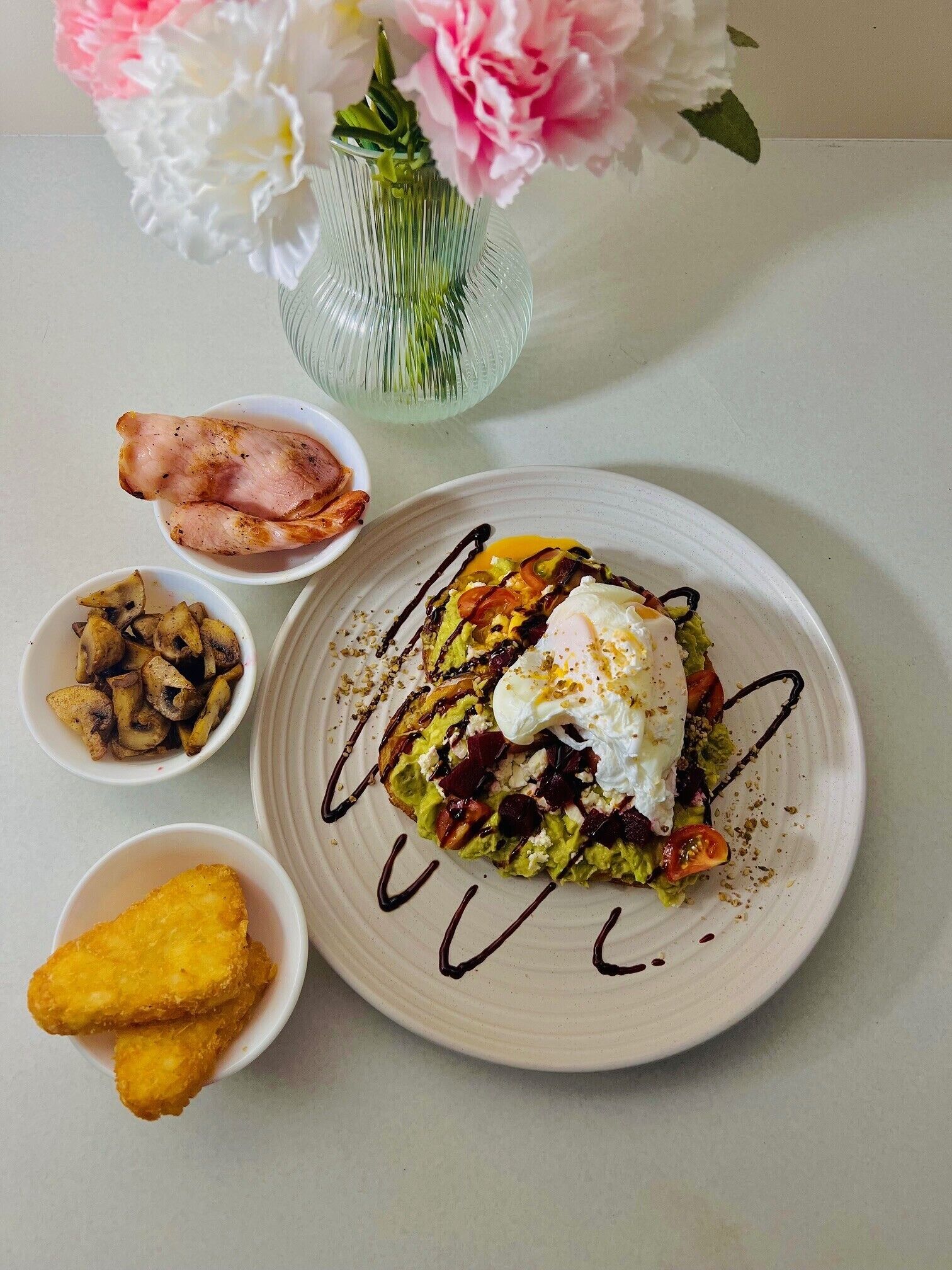 A plate of food with a poached egg, avocado, and red onions, accompanied by a side of fried potatoes and a small bowl of bacon.