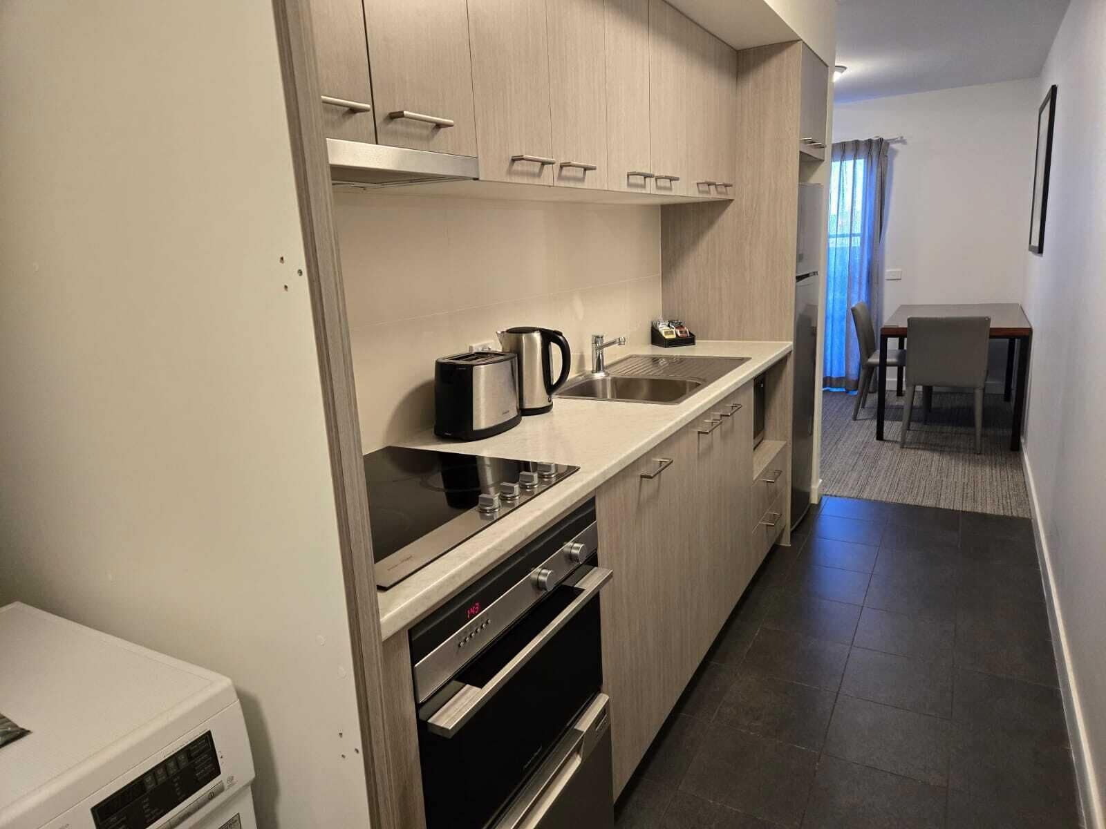 A modern kitchen with a built-in oven, a stovetop, and a sink. There is a dining area with a table and chairs visible in the background.