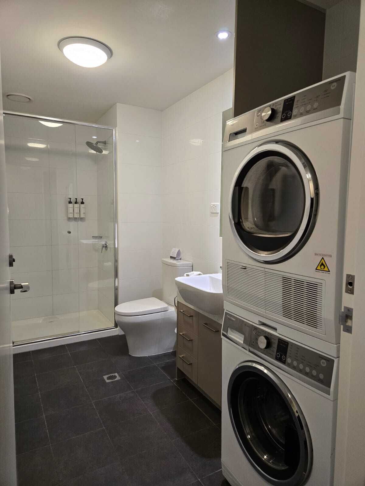 A modern bathroom with a glass shower enclosure, a toilet, a sink, and stacked laundry appliances.