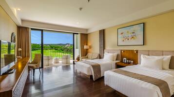 Habitación de hotel con dos camas, un escritorio con televisor y un balcón con vistas a la vegetación.