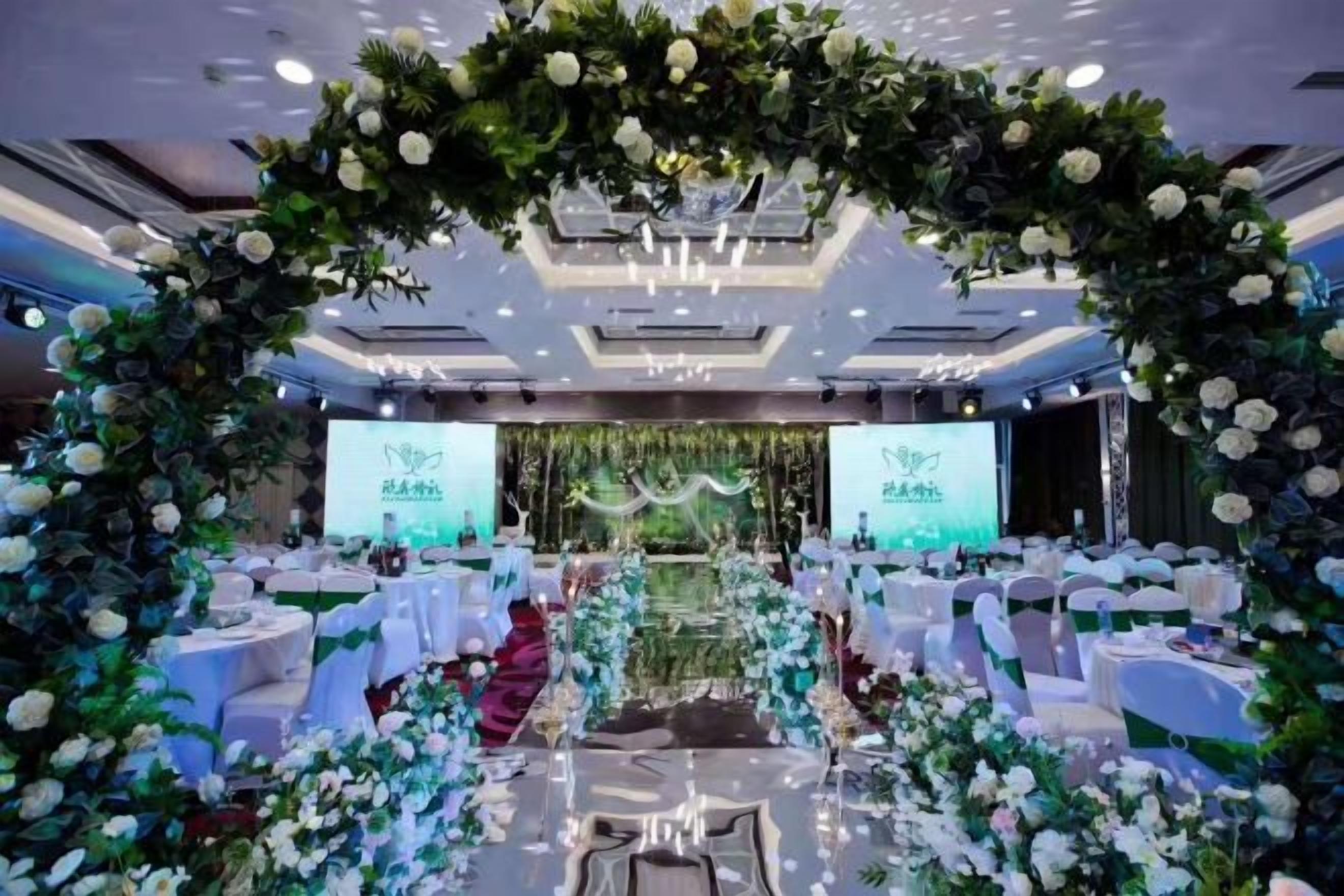 Un salón de banquetes con mesas redondas, un escenario con fondo floral e iluminación empotrada en el techo.