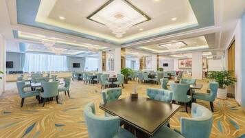 A spacious hotel dining area with multiple tables and chairs, a patterned carpet, and a modern ceiling design.