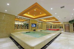 A large indoor swimming pool with a tiled floor, a ceiling with recessed lighting, and a television screen mounted on the wall.