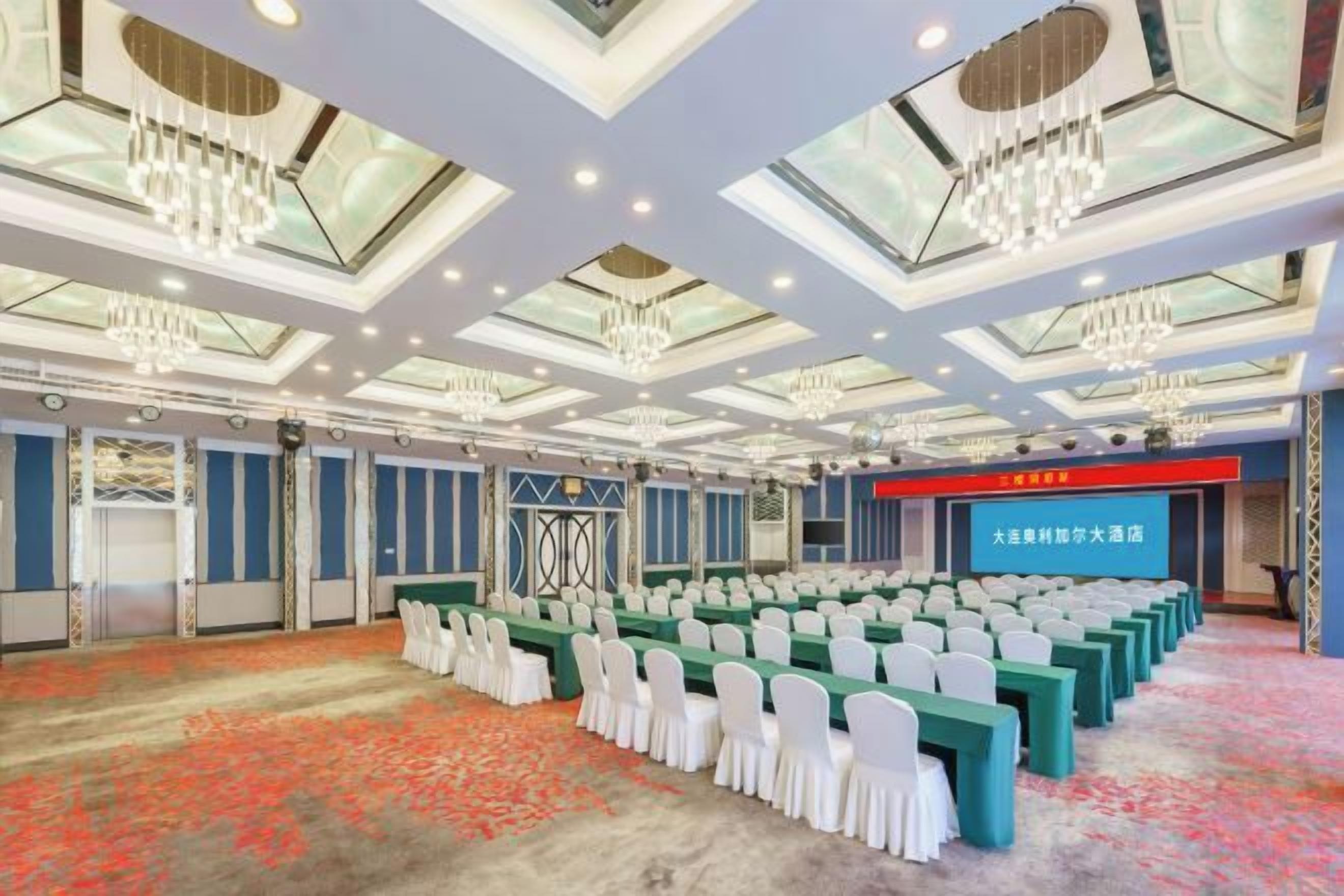 A conference room with rows of tables and chairs, a stage, and a large screen.
