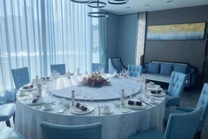 A round dining table set for six with chairs, a sofa, and a large window with curtains. - Aulicare Hotel (Dalian)
